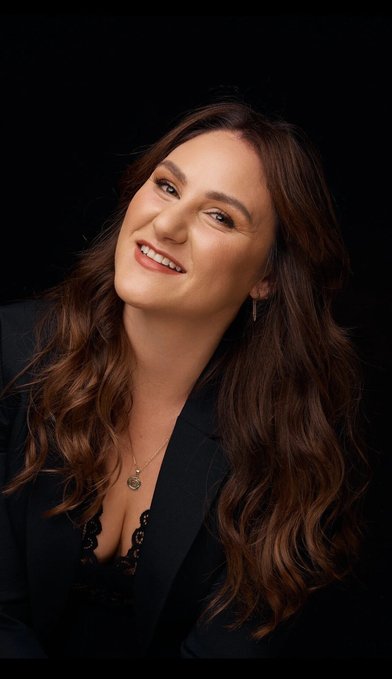 A woman with long wavy brown hair smiling and winking at the camera, wearing a black blazer and a necklace against a black background.
