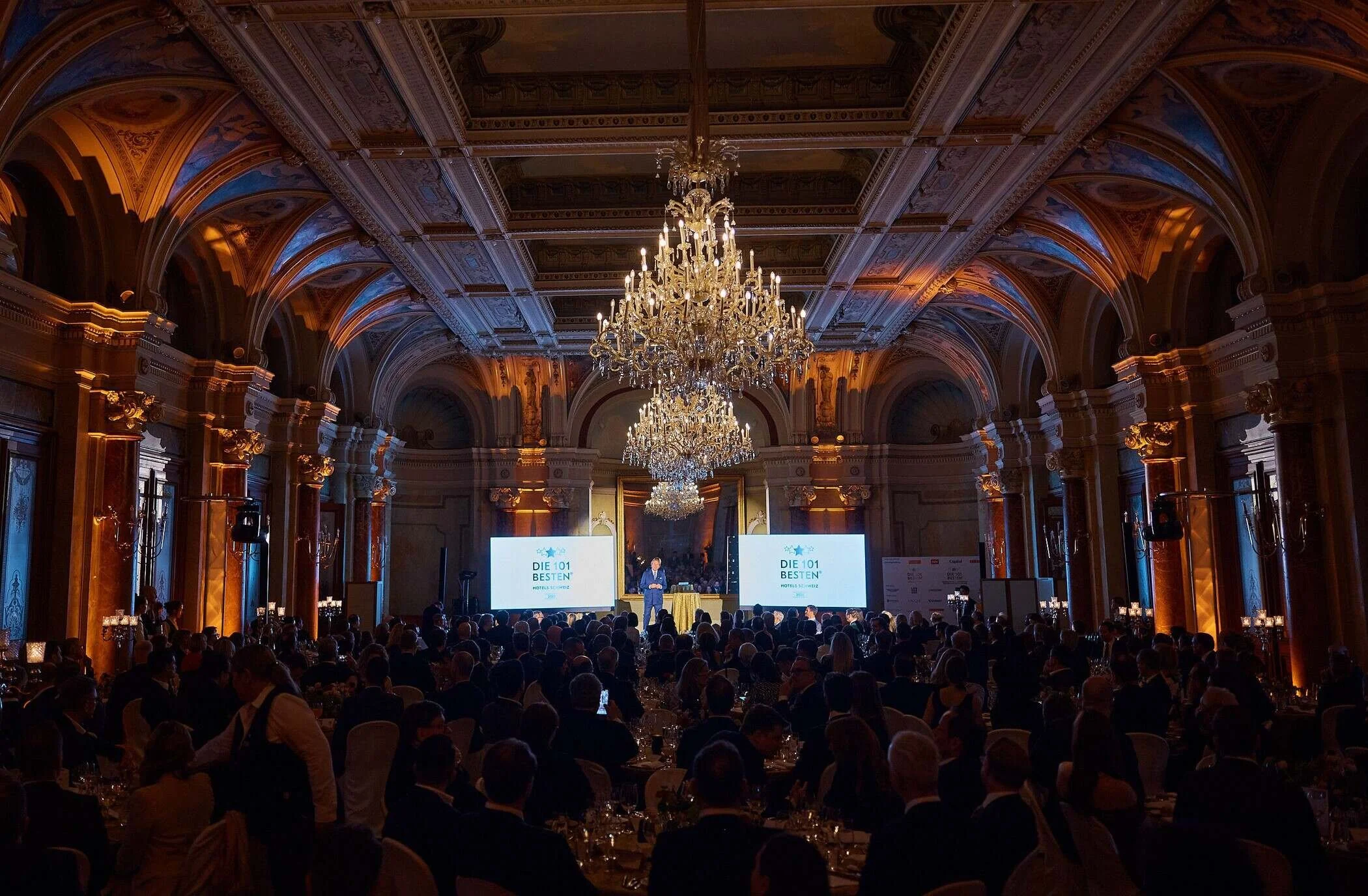 Elegant_banquet_hall_with_chandeliers_and_audience.jpg
