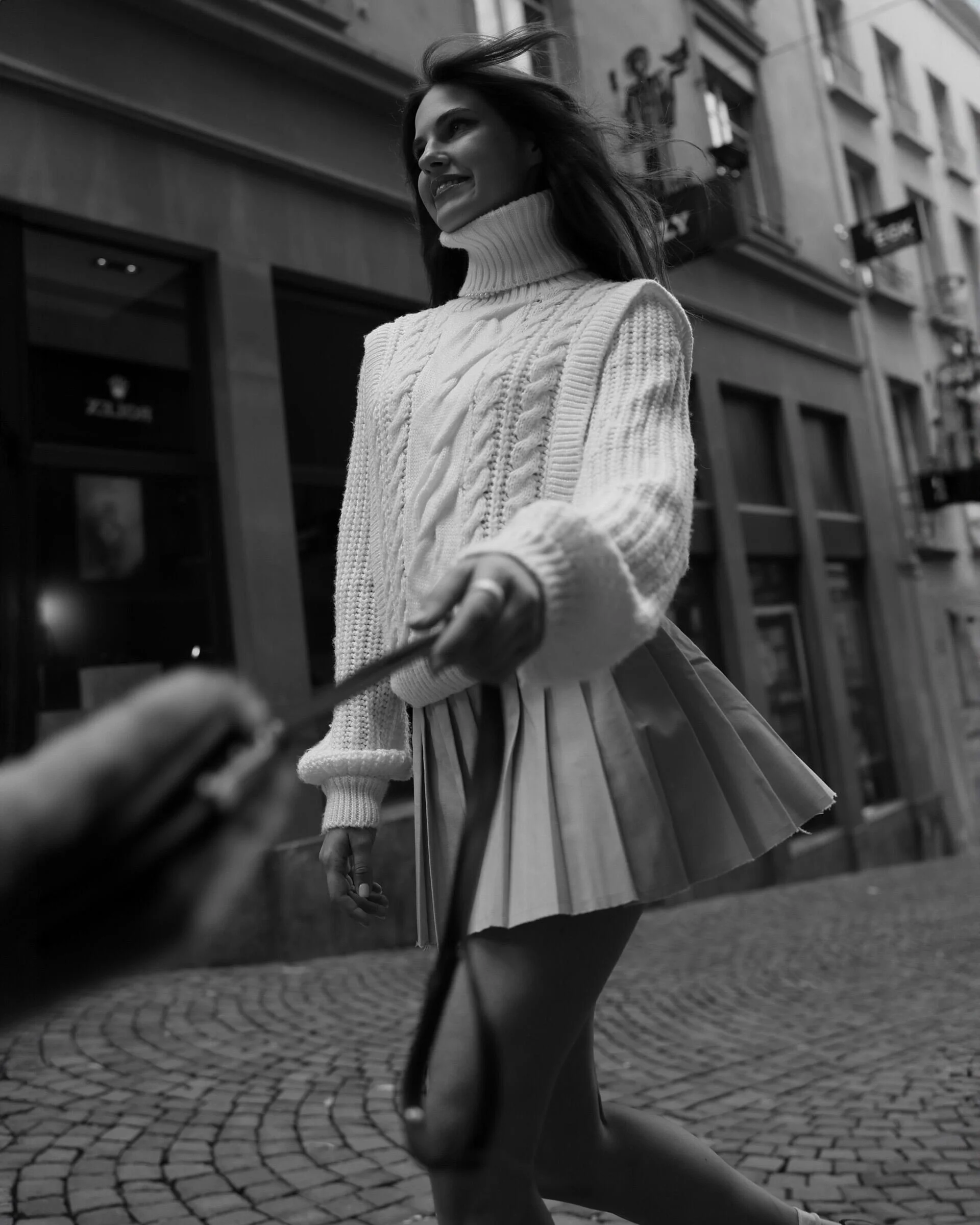 Woman_in_white_sweater_and_pleated_skirt_walking_on_cobblestone_street.jpg