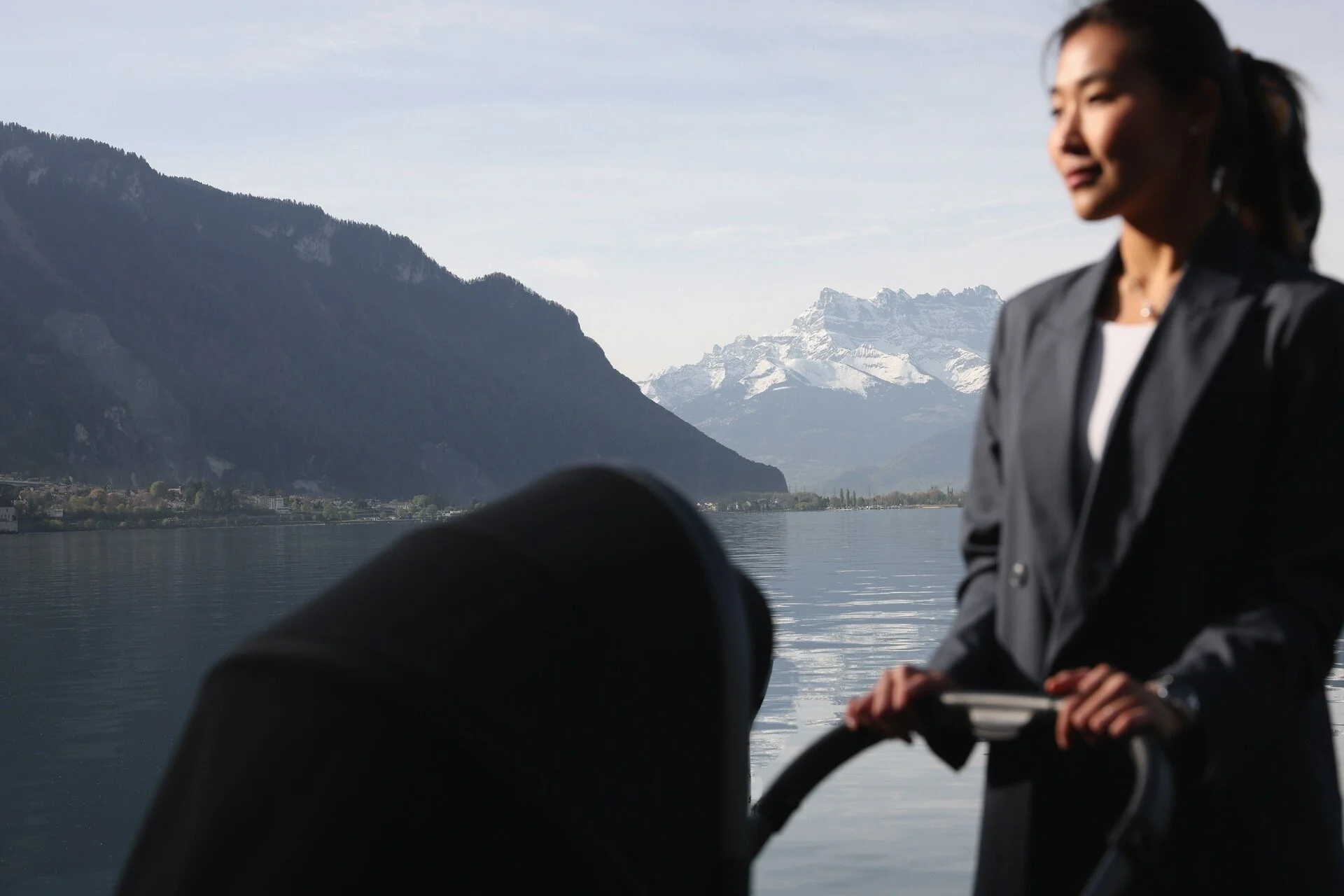 Une femme vêtue d'un manteau gris se tient au bord d'un lac, avec des montagnes en arrière-plan. Cette photo a été prise à Montreux par un photographe professionnel.