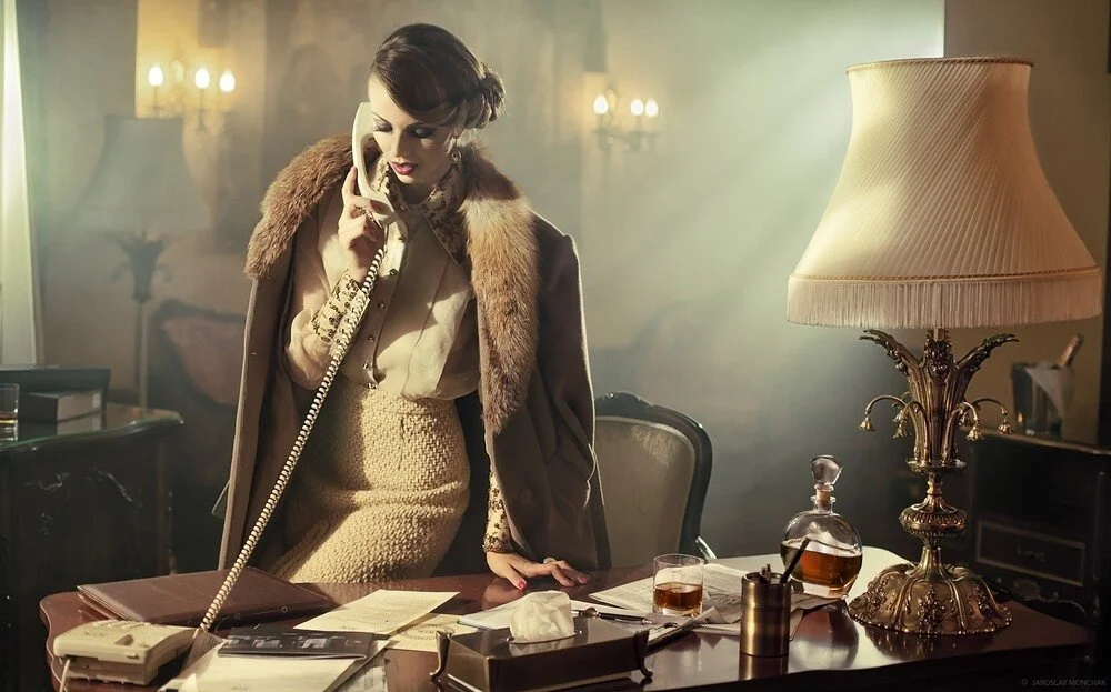 Woman_in_vintage_attire_at_a_desk_with_a_phone_and_lamp.jpg