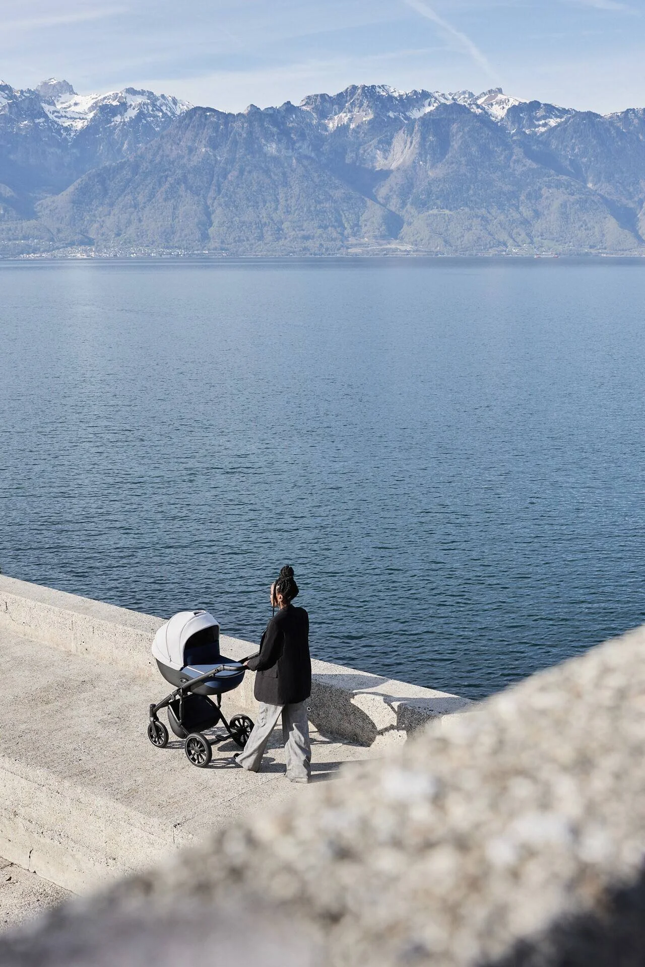 Une femme vêtue d'un manteau noir et d'un pantalon gris pousse une poussette le long d'un sentier au bord du lac, avec des montagnes en arrière-plan. Capturée à Montreux par un photographe professionnel.