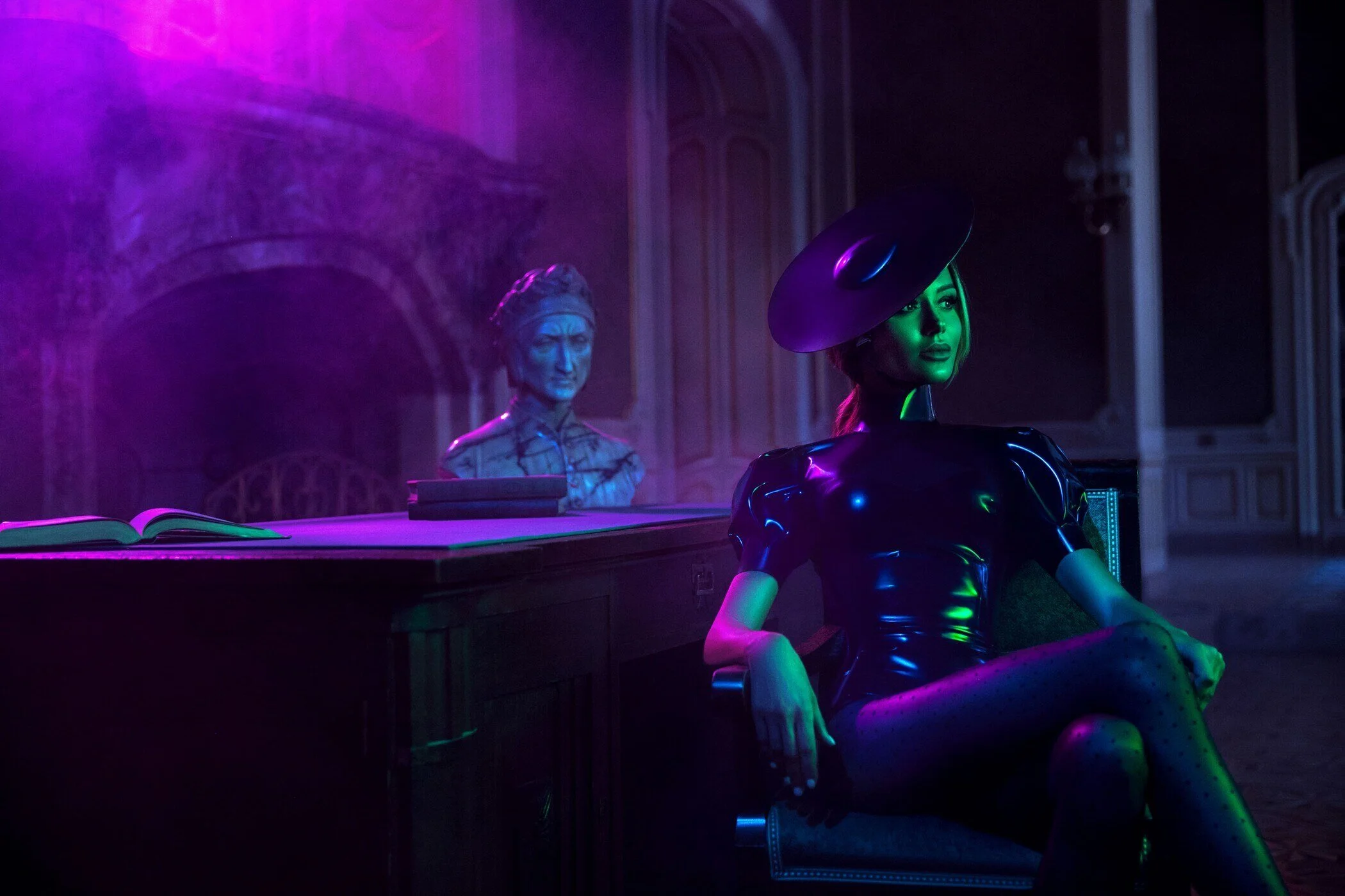 Une femme assise sur une chaise, vêtue d'une tenue noire brillante et d'un grand chapeau, entourée d'un éclairage violet et vert. Capturée à Genève par un photographe portraitiste professionnel.