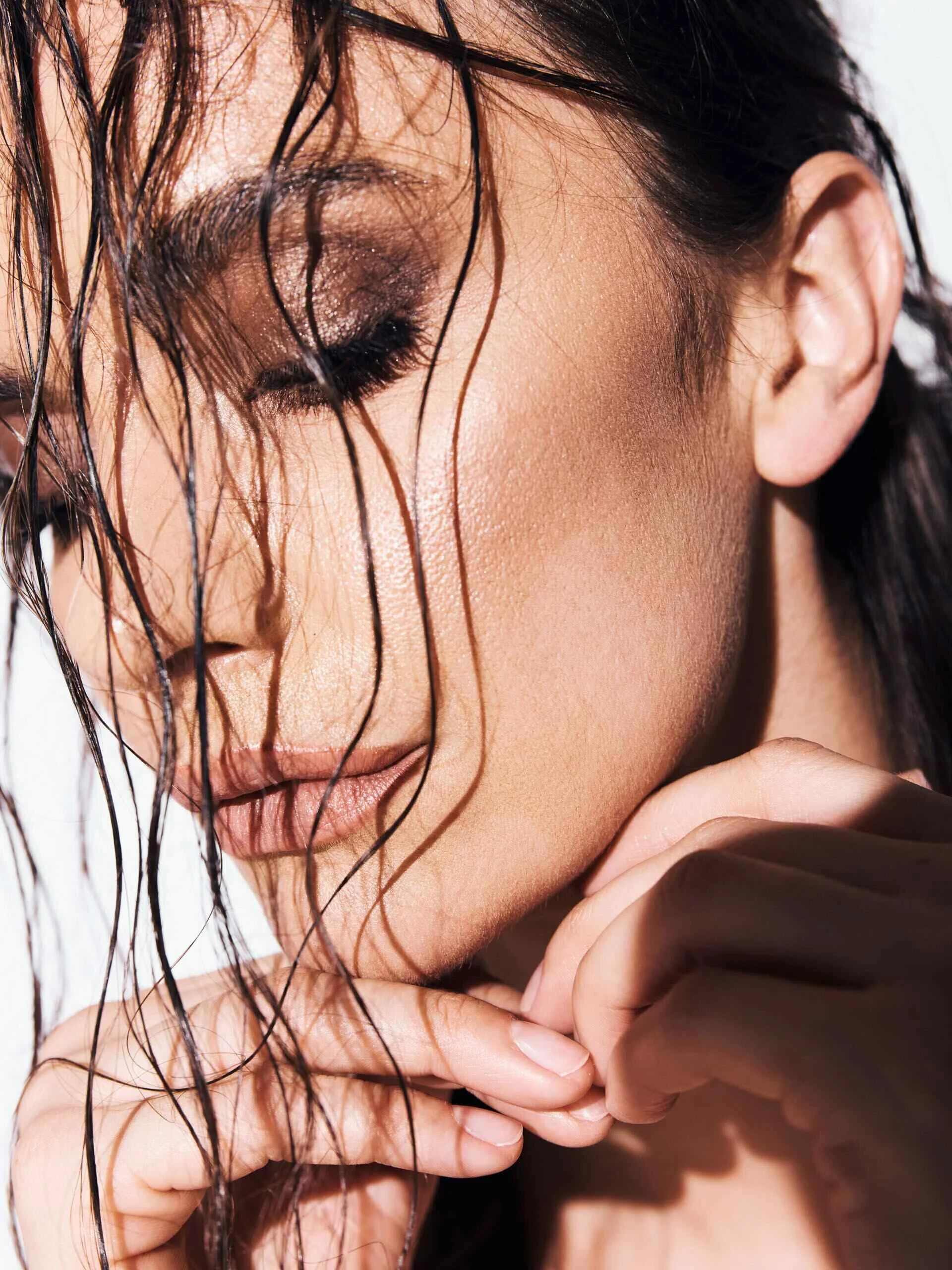 Close-up of a woman with dark wet hair, partially wet face, wearing shimmery eyeshadow, with her eyes closed, and hand near her chin.