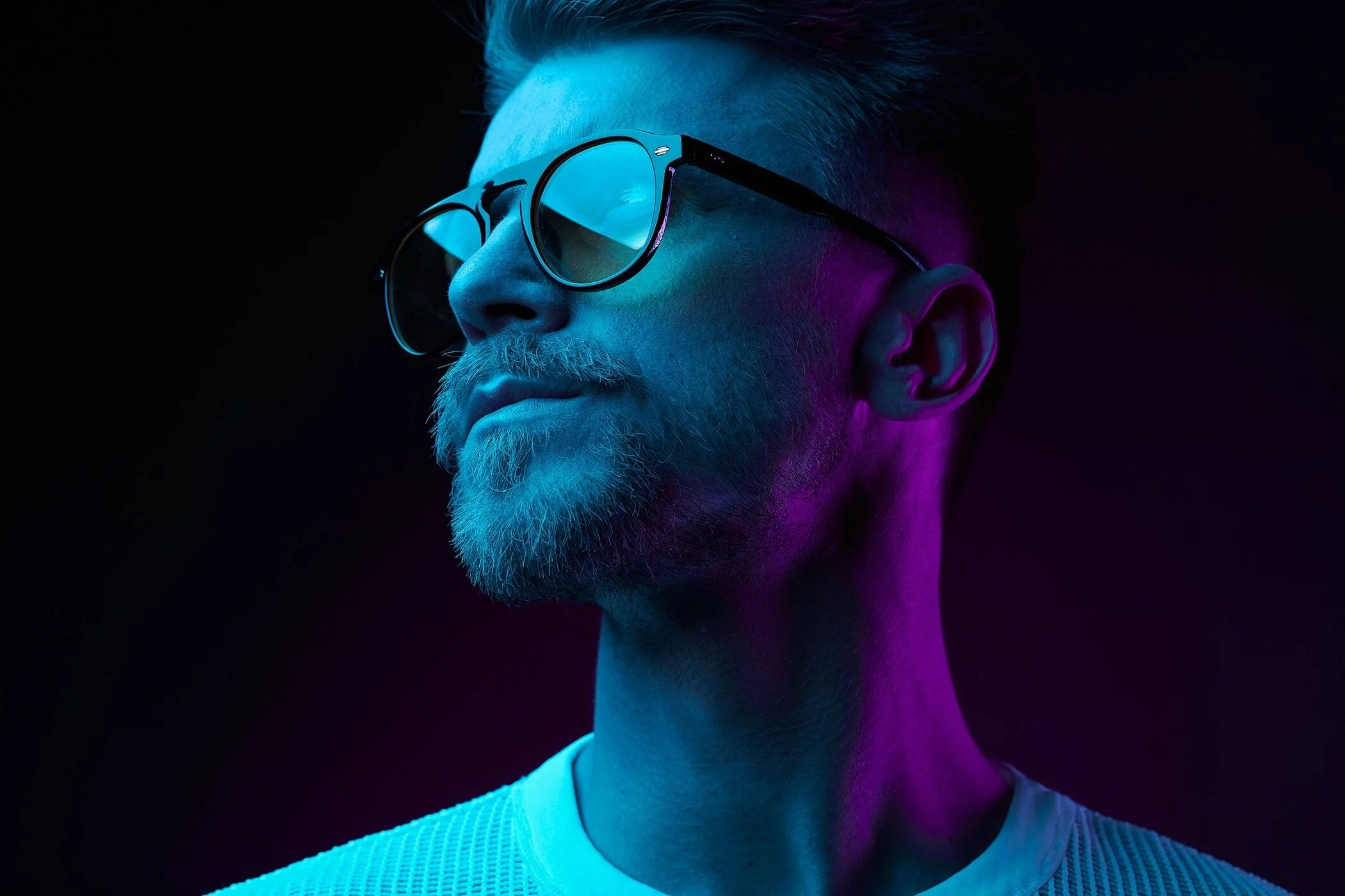 L'image représente un homme portant des lunettes de soleil, éclairé par des lumières bleues et violettes sur un fond sombre. Elle a été prise à Zurich par un photographe professionnel.