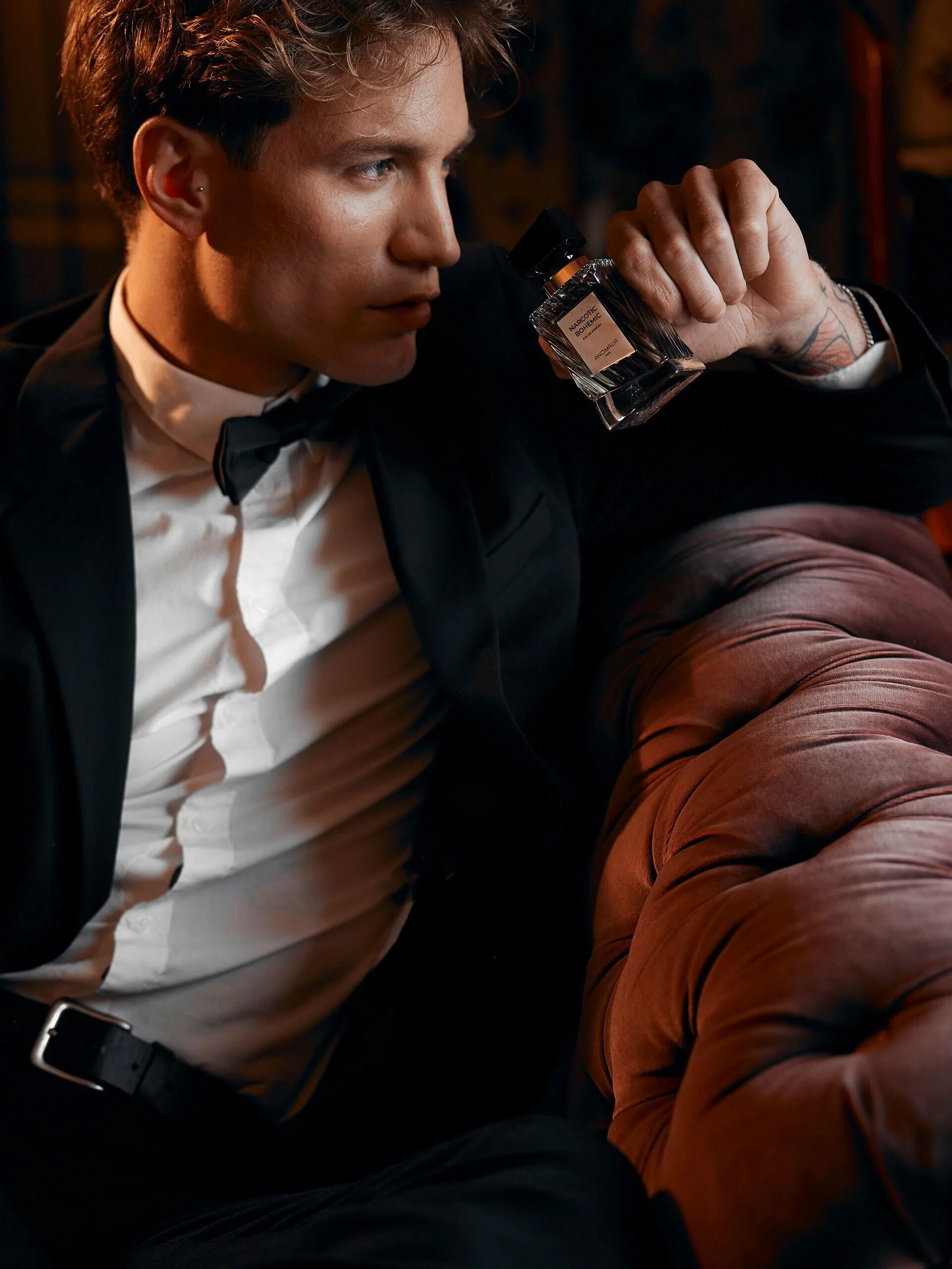 Un homme en smoking tient un flacon de parfum assis sur un canapé en velours. Capturé à Zürich par un photographe portraitiste professionnel.