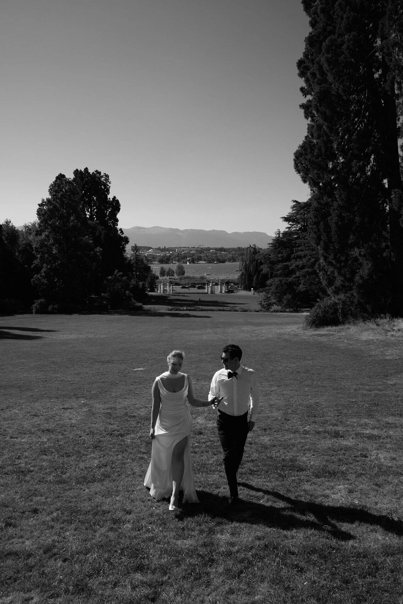 Une photo en noir et blanc représentant une mariée et un marié marchant main dans la main sur l'herbe, avec de grands arbres et des montagnes en arrière-plan.