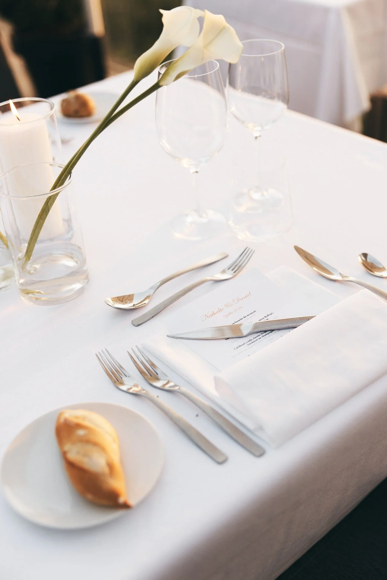 Une table à manger habillée, avec trois verres à vin vides, une bougie, un petit bouquet de callas et des couverts dorés posés sur une nappe blanche.