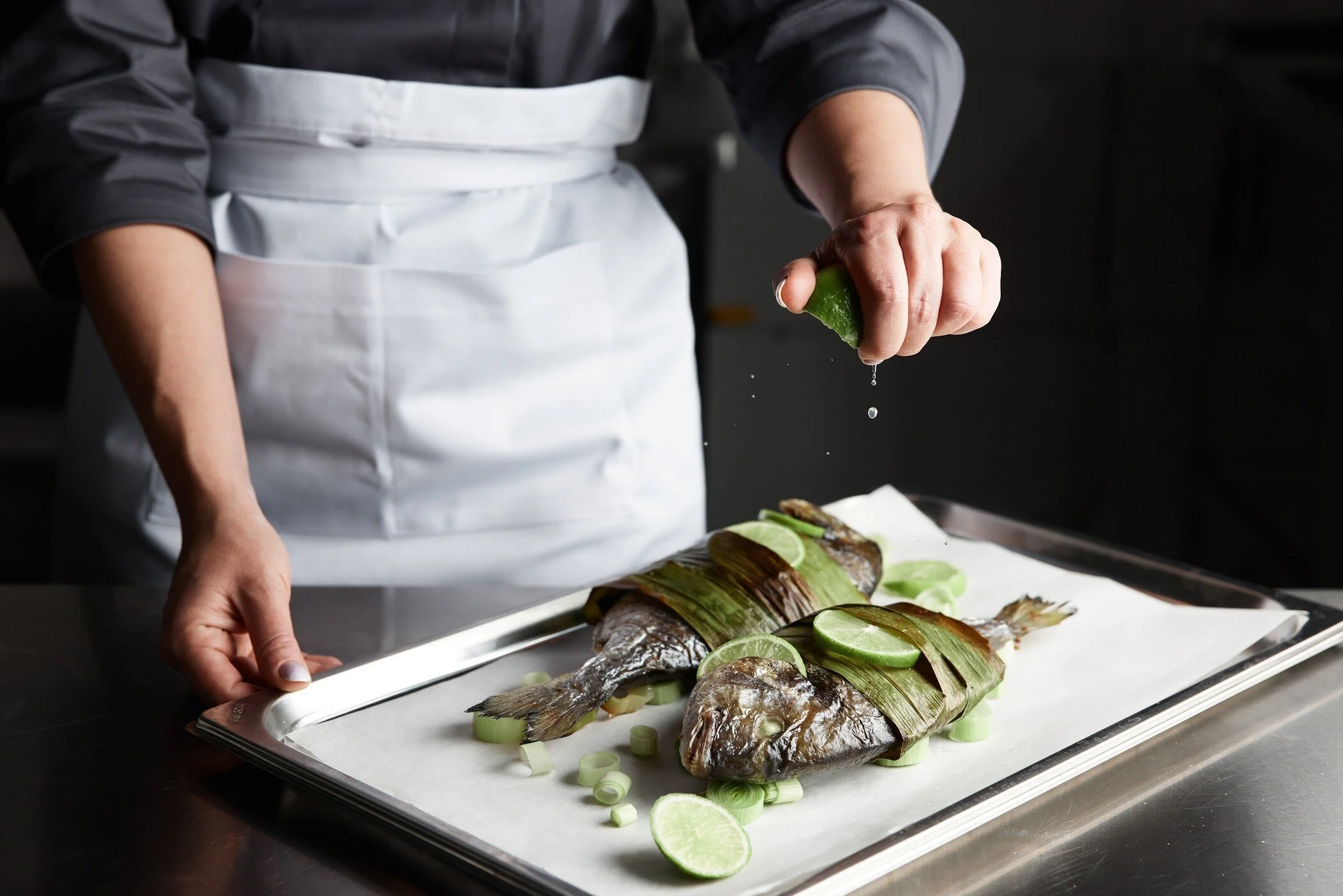 A chef in a dark shirt and white apron squeezes lime over two fish on a tray, garnished with green onions and lime slices. Captured in Switzerland by a professional portrait photographer.