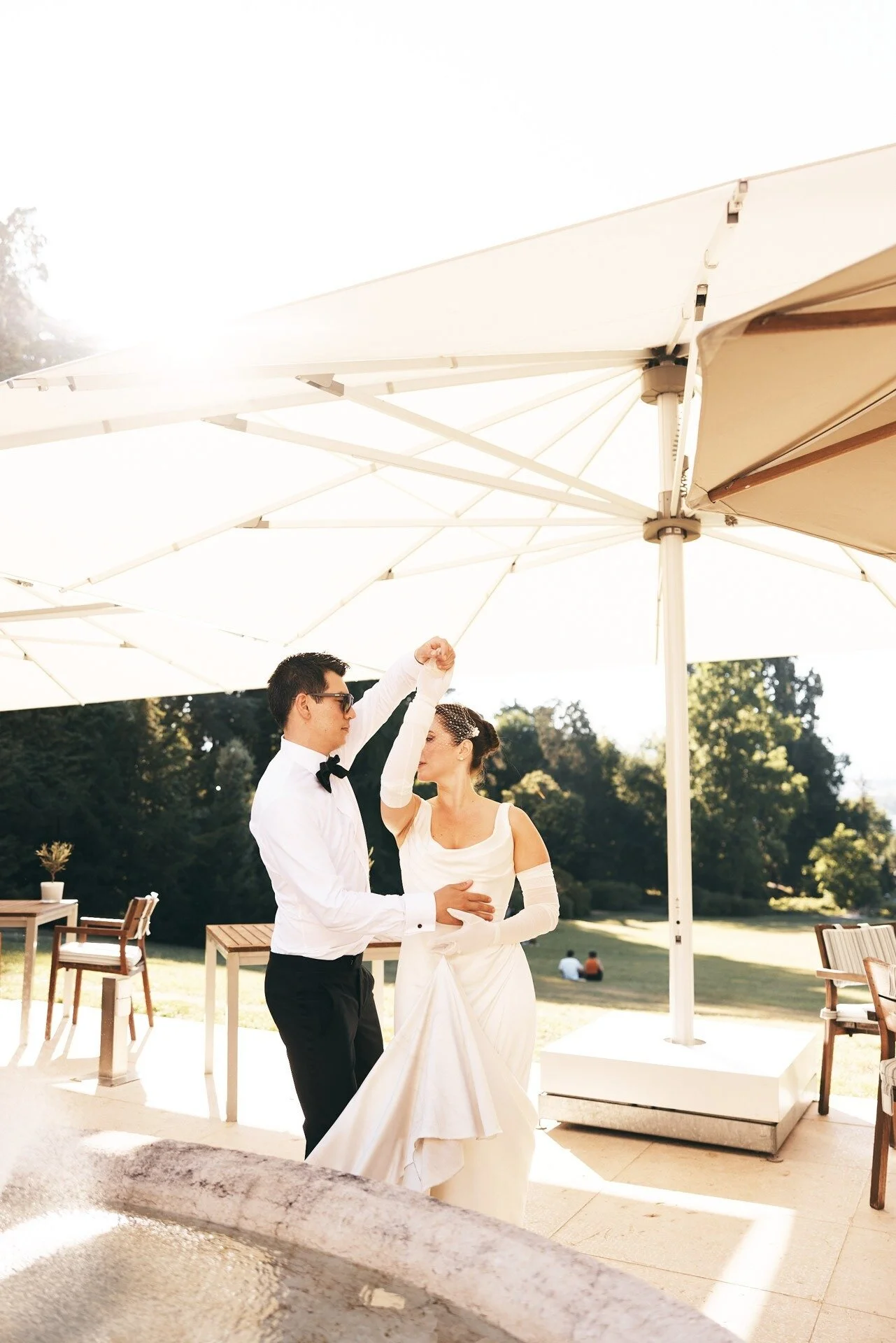 Un couple danse en plein air sous un grand parasol blanc, avec des arbres et une pelouse en arrière-plan. L'homme porte un smoking et des lunettes de soleil, tandis que la femme est vêtue d'une robe blanche.