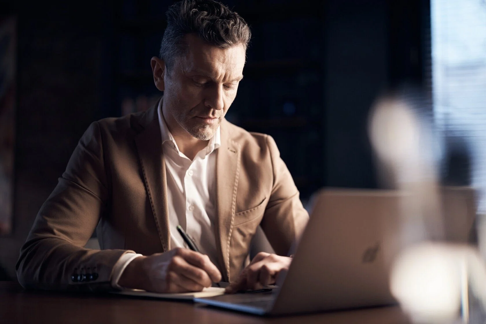 Man_in_brown_blazer_writing_at_desk_with_laptop.jpeg