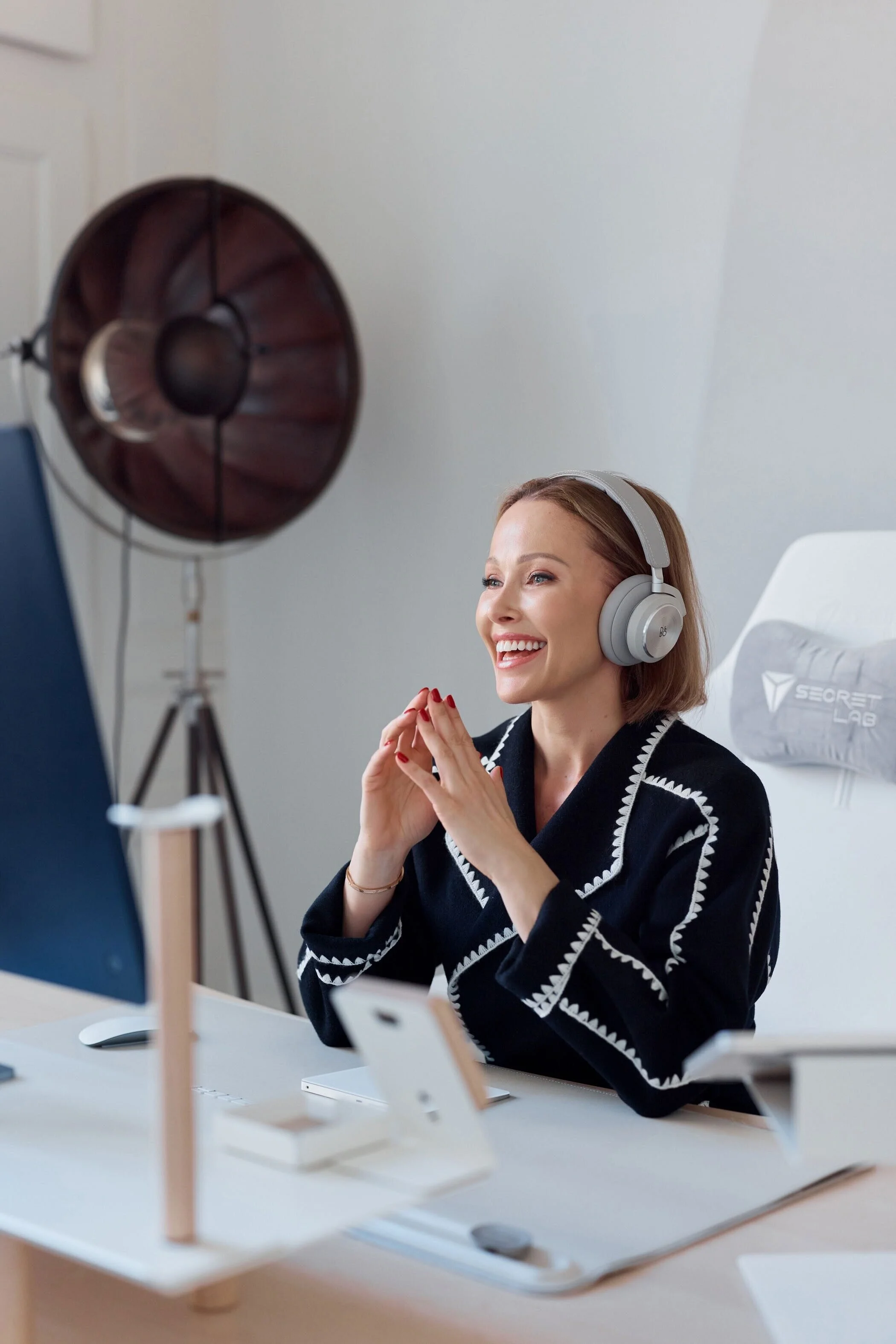 A woman with light brown hair and red lipstick, wearing a black sweater with white embroidered edges, smiling and sitting at a desk with a computer and smartphone, wearing gray over-ear headphones, in a well-lit room with a large studio light on a st
