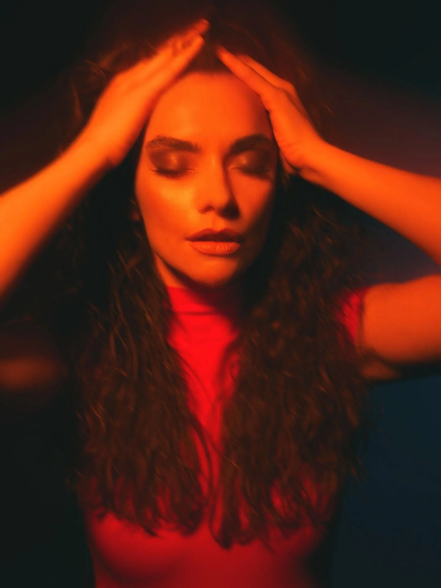 A woman with curly hair and makeup, eyes closed, with her hands on her head.