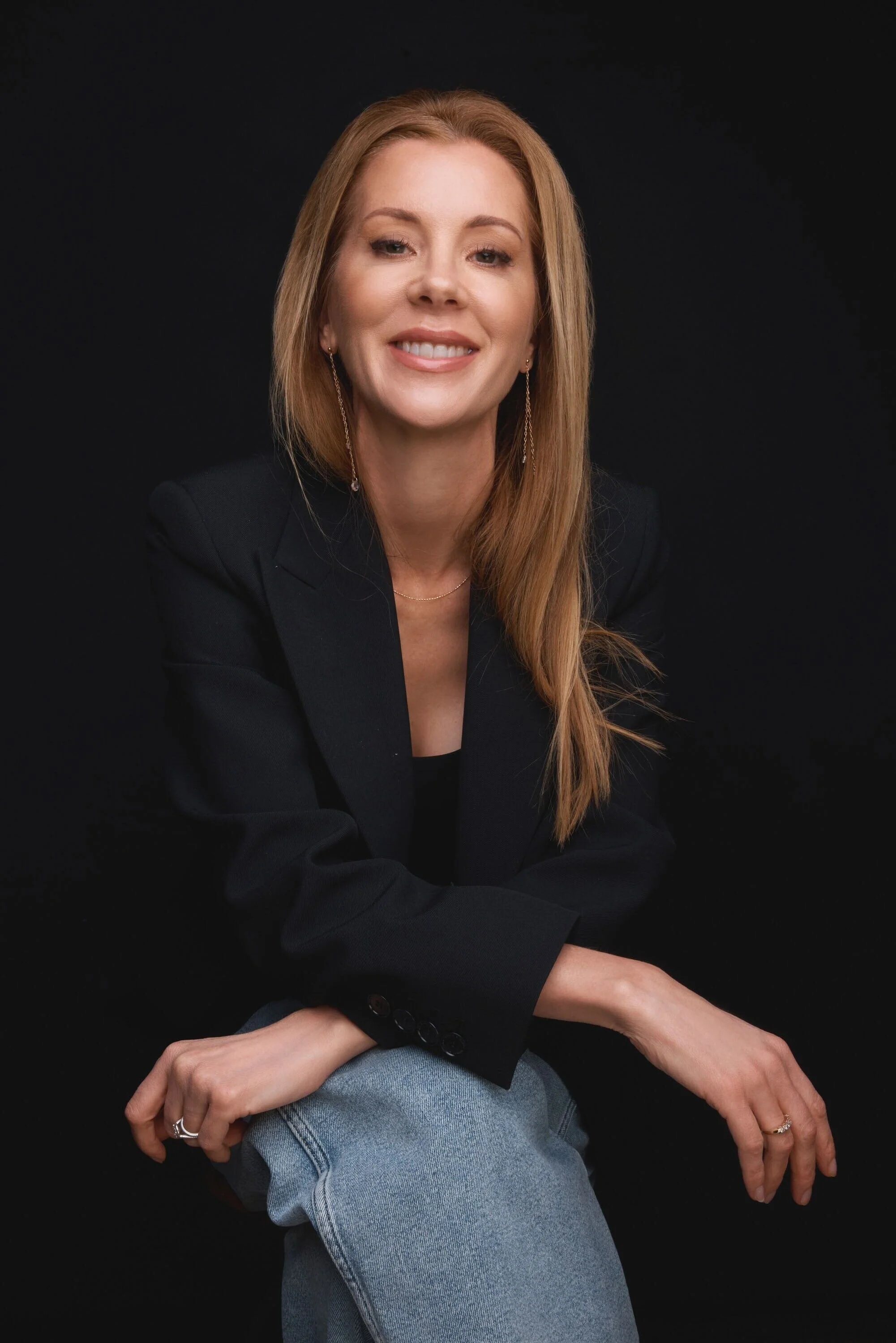 A woman with long, wavy red hair smiling in front of a black background. She is wearing a black blazer, light blue jeans, and gold jewelry.
