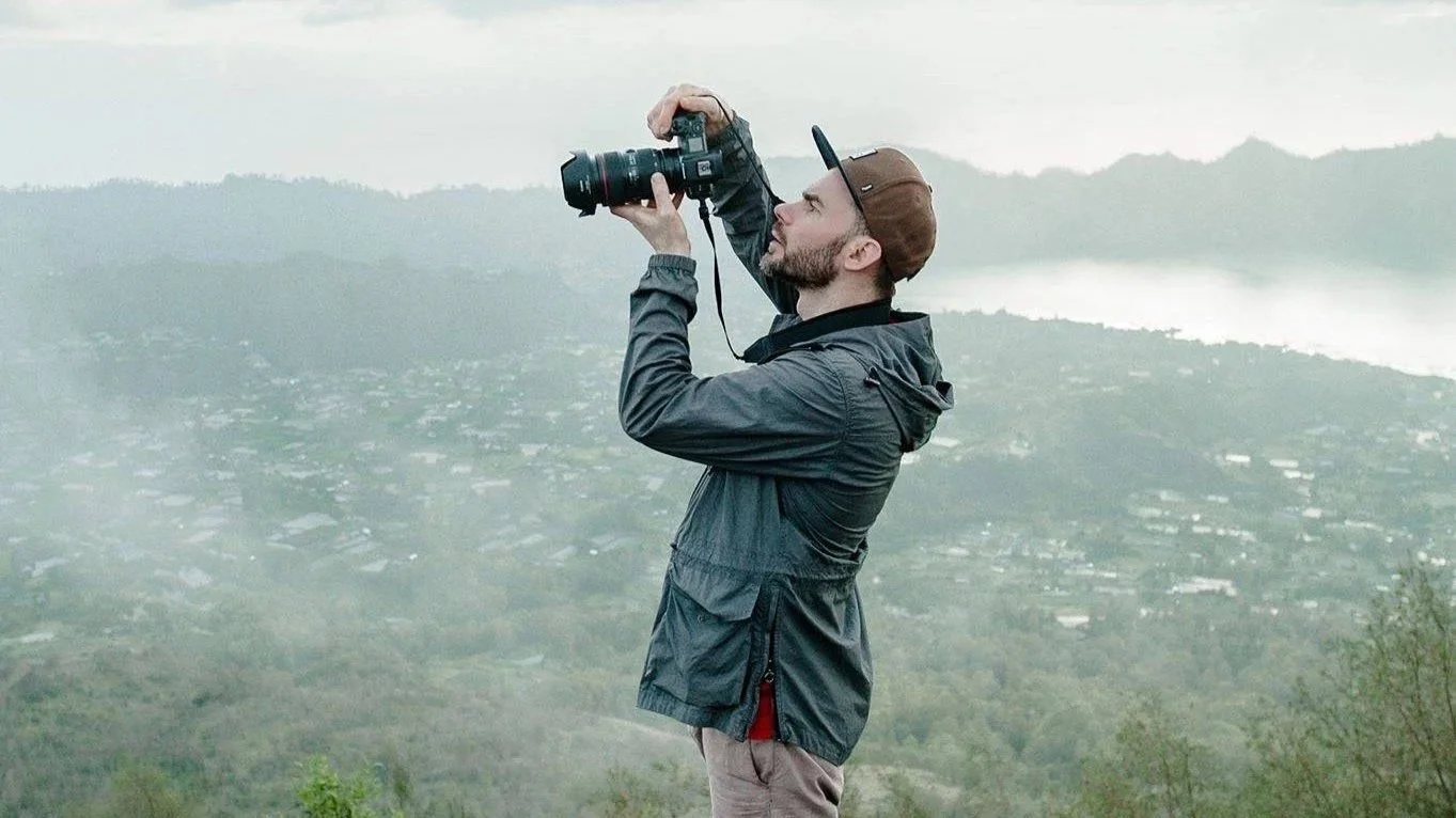 A man in a grey jacket and brown cap stands on rocky terrain, photographing a distant landscape. Captured in Montreux by a professional portrait photographer.