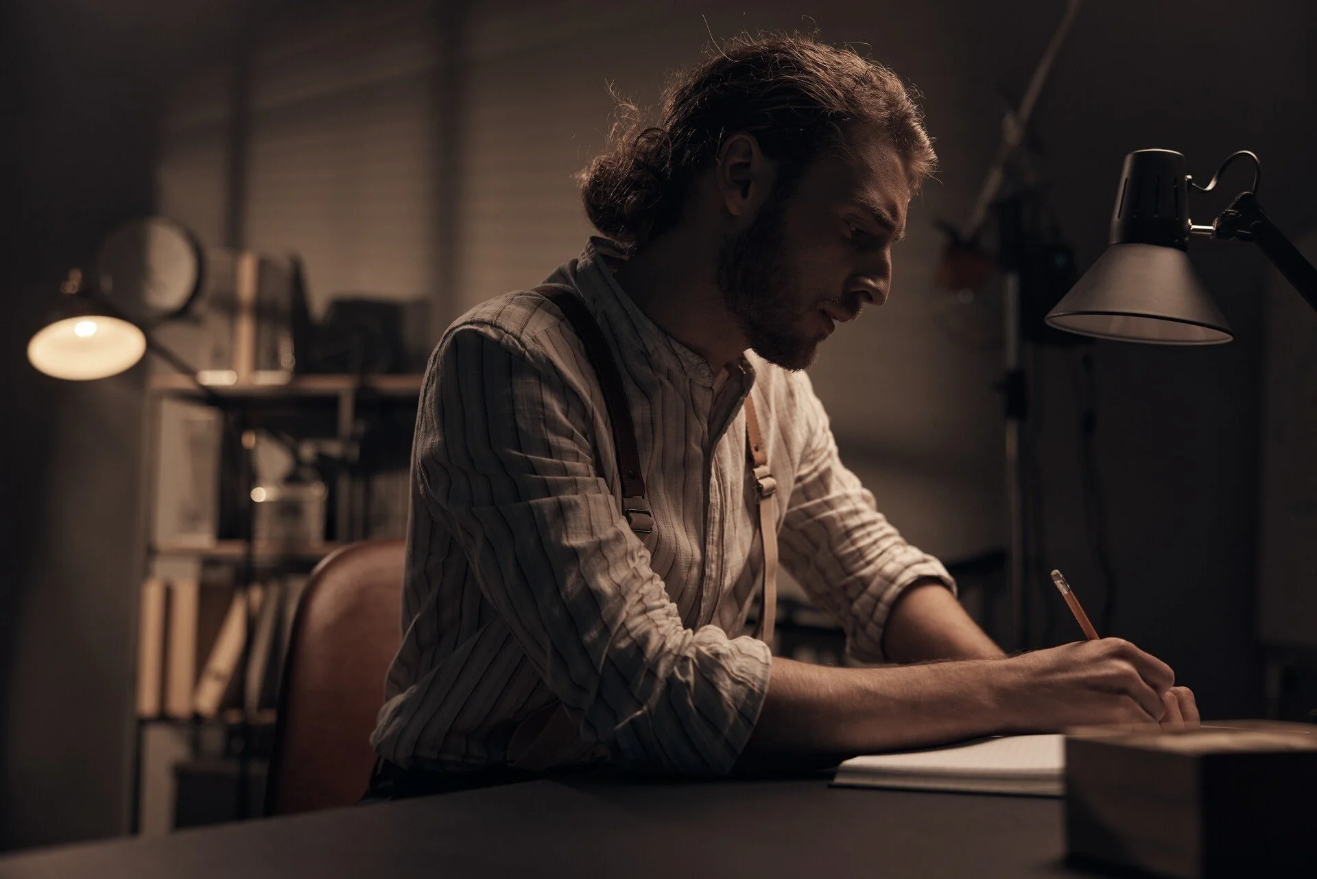 Un homme aux cheveux longs et aux bretelles écrit à un bureau sous une lampe. Capturé à Zürich par un photographe portraitiste professionnel.