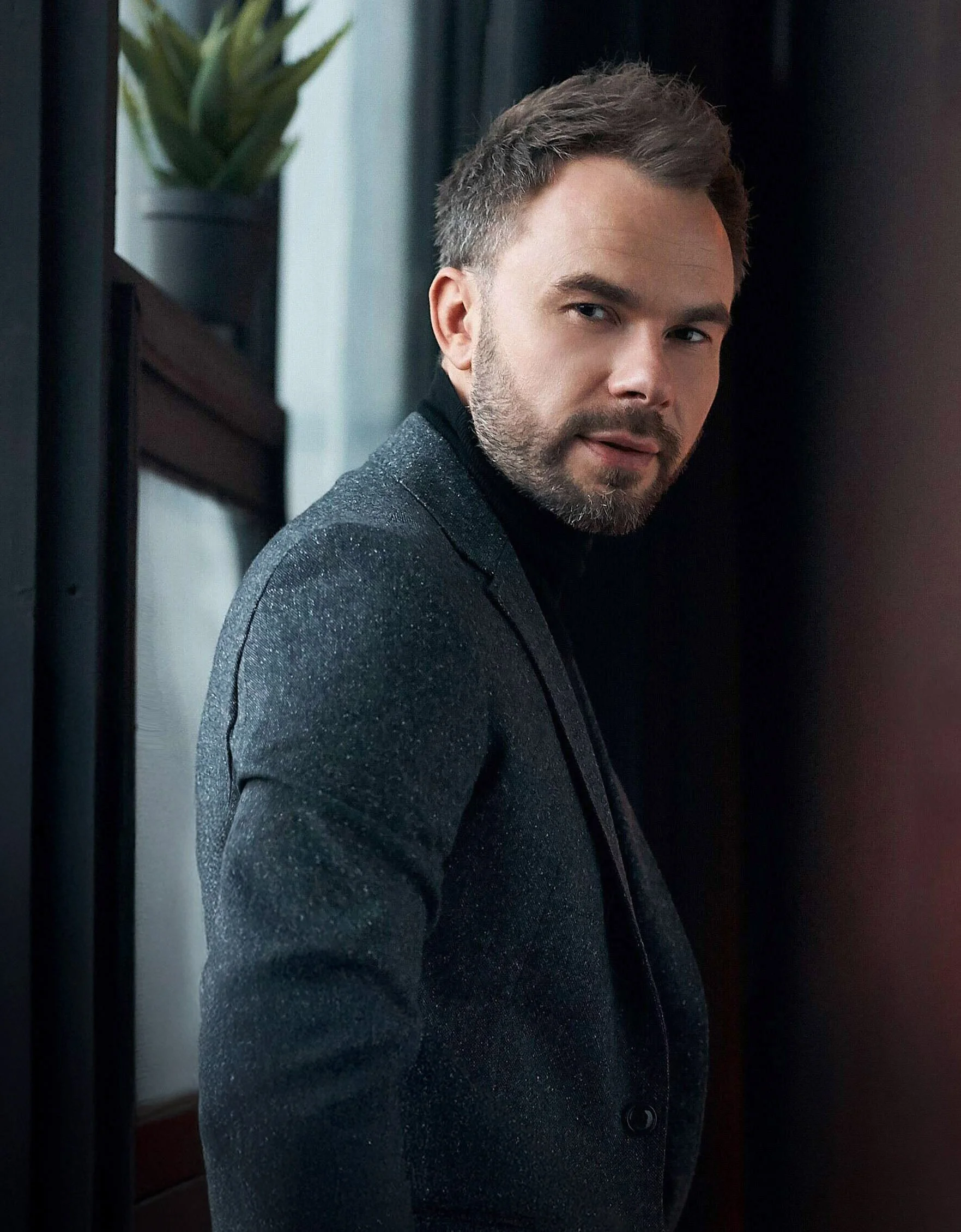 Yaroslav Monchak, vêtu d'un col roulé noir et d'un blazer sombre et scintillant, debout à l'intérieur, près d'un mur noir et d'une plante.