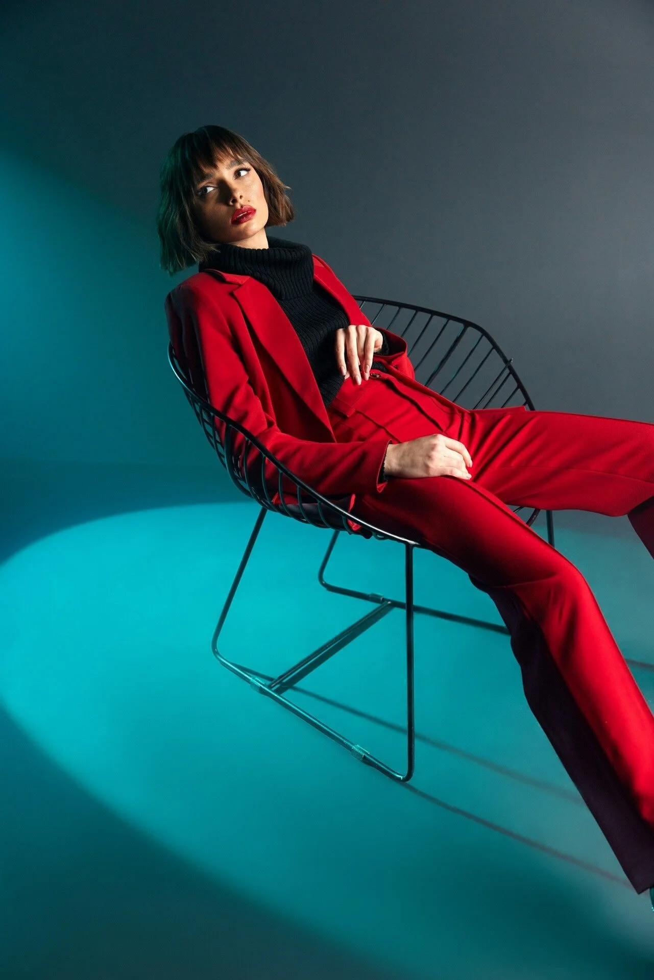 Une femme en tailleur rouge et col roulé noir est assise sur une chaise en fil de fer, éclairée par une lumière sarcelle. Cette photo a été prise à Bâle par un photographe professionnel.