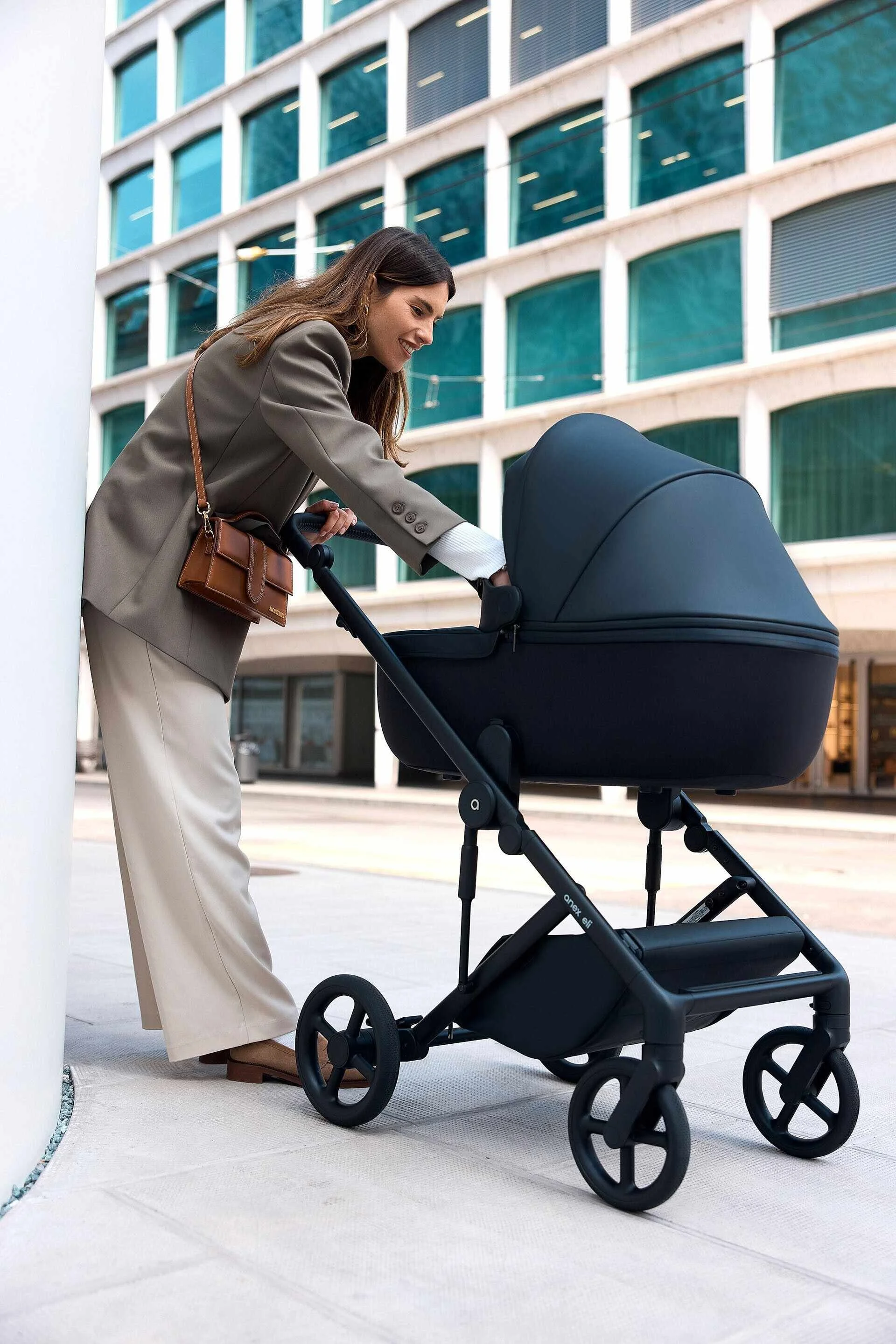 Une femme en tailleur beige se penche sur une poussette noire dans un environnement urbain moderne. Capturé à Genève par un photographe professionnel.