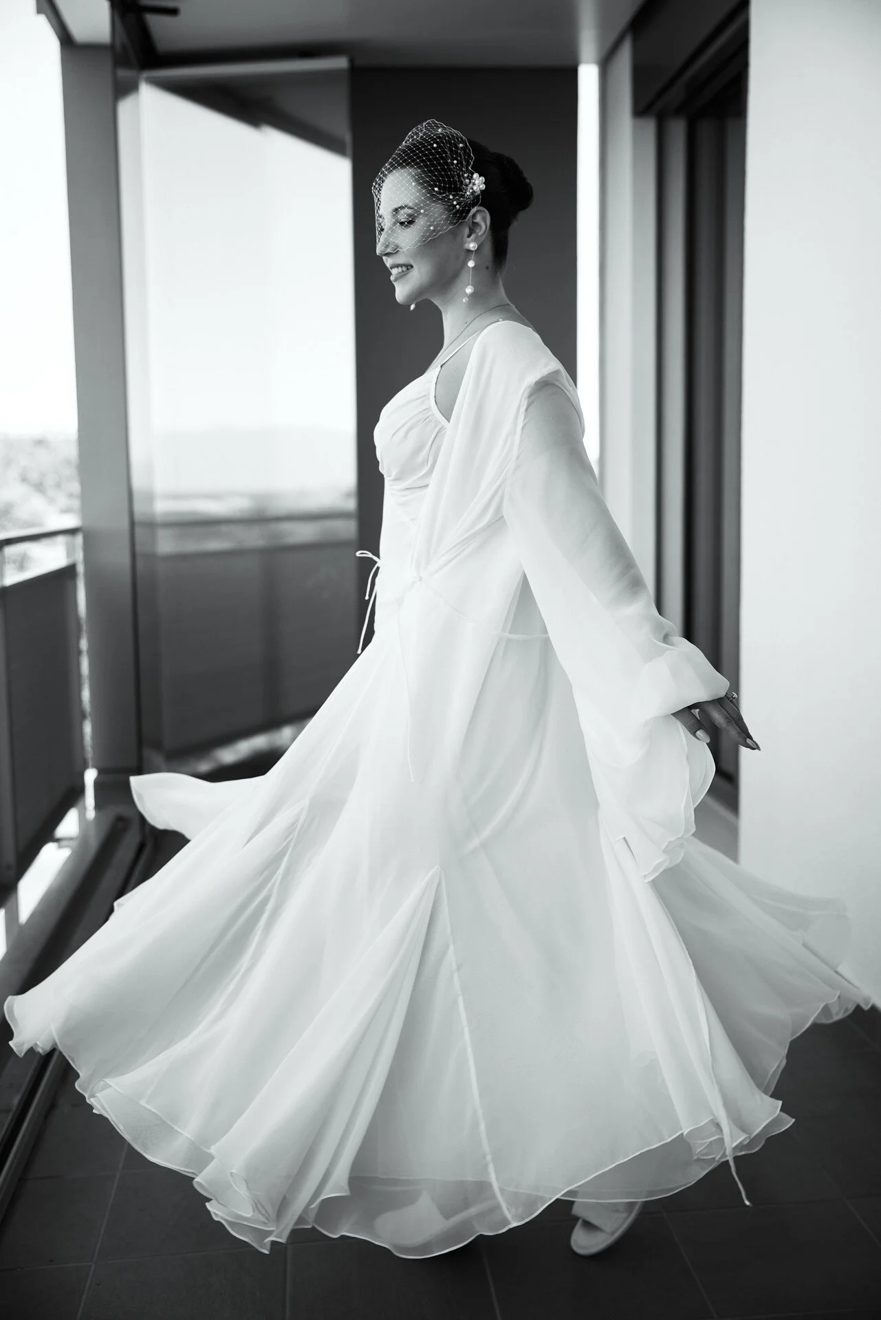 Une femme vêtue d'une robe de mariée blanche et portant des boucles d'oreilles en perles, ainsi qu'un voile cage, marche dans un décor intérieur moderne.