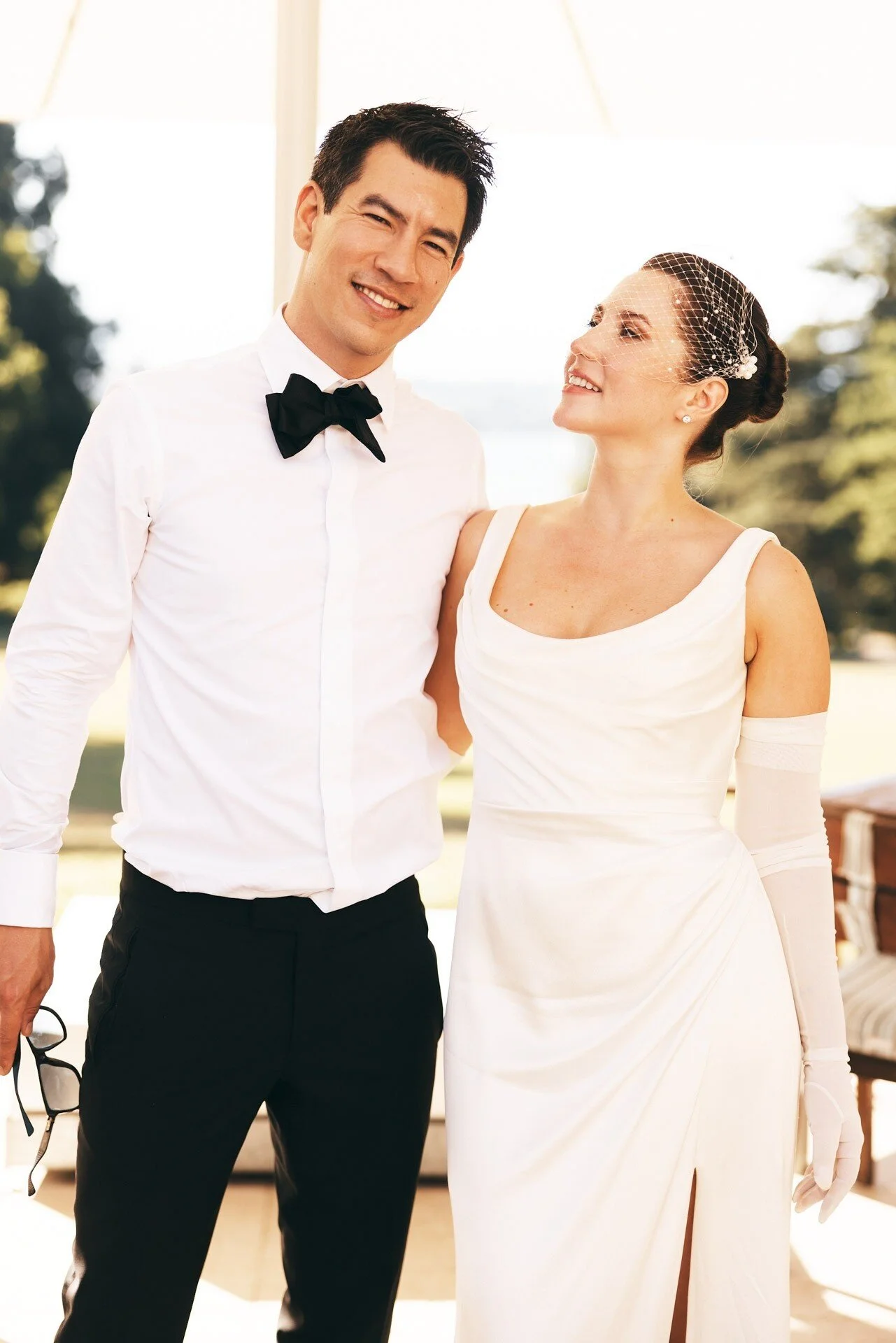 Un homme et une femme en tenue de mariage, debout à l'extérieur, souriants, la femme regardant l'homme. L'homme porte une chemise blanche, un nœud papillon noir et un pantalon noir ; il tient un verre à la main ; la femme porte une robe blanche fendue et un bi