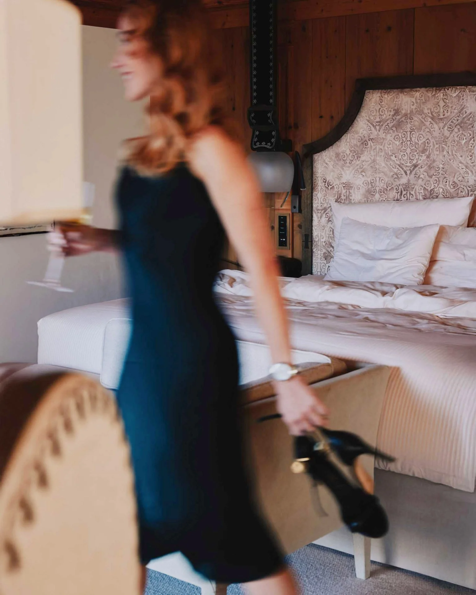 A woman in a black dress walks through a hotel room holding black shoes. Captured in Switzerland by a professional portrait photographer.