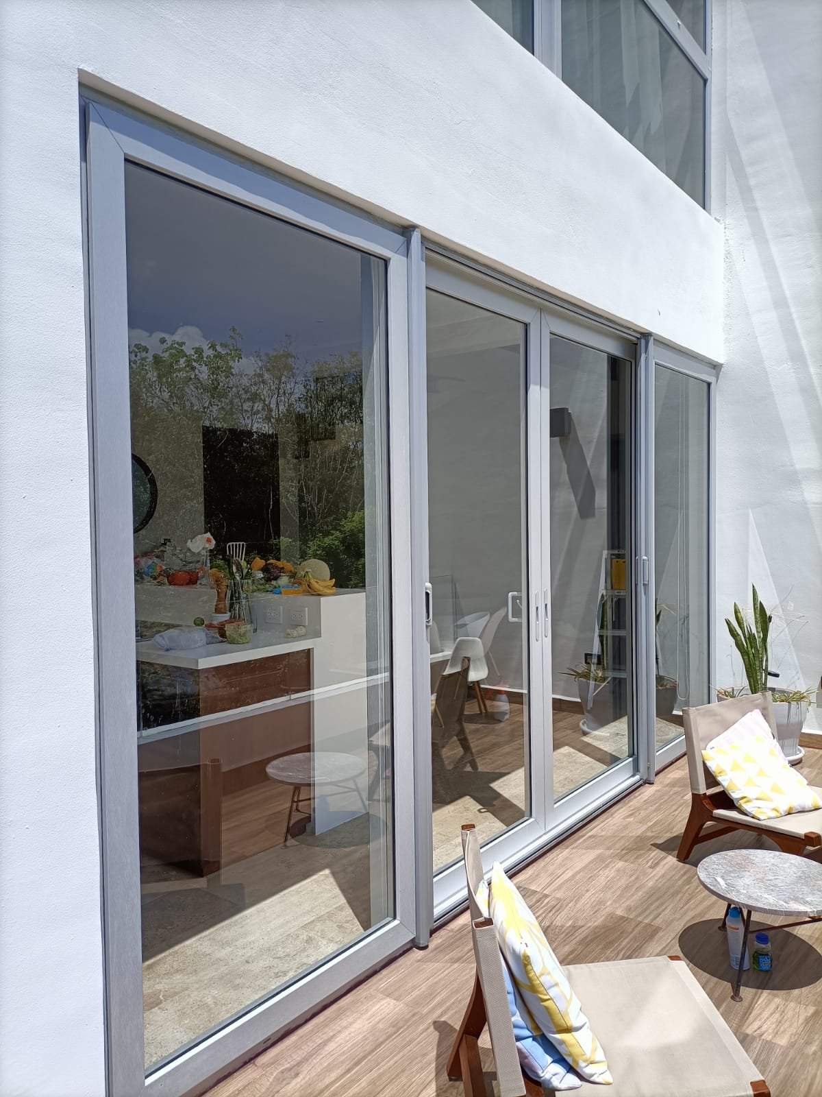 Modern home exterior with large glass sliding doors leading to an outdoor patio area with table and chairs, potted plants, and reflection of trees in the glass.