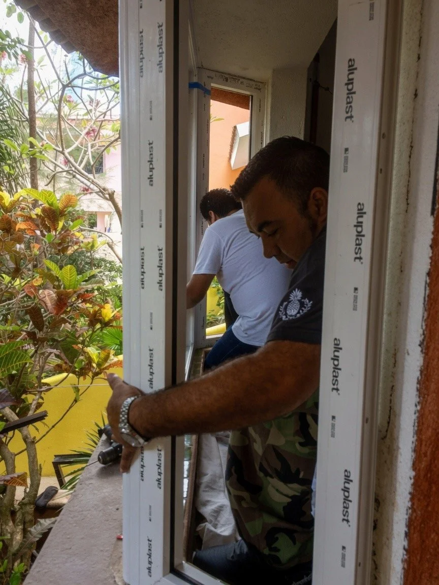 Men installing a window frame on a balcony, with one man holding the frame and another working nearby, surrounded by plants and residential buildings.