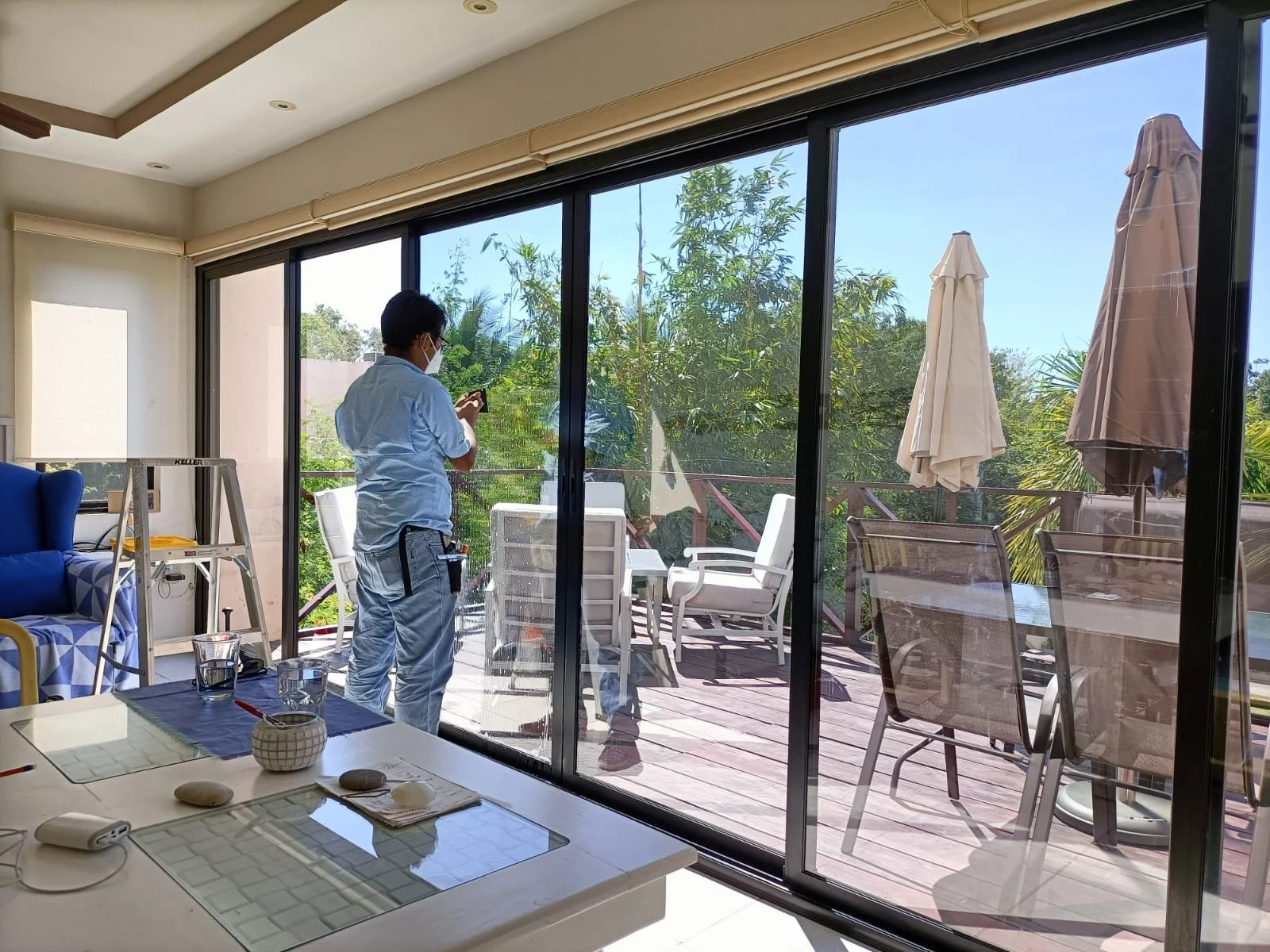 A man wearing a blue shirt and white pants, with a tool belt, stands on a deck outside, looking at a smartphone, with patio furniture and umbrellas around. Inside, the room has a table with glassware and kitchen items, and large sliding glass doors leading to the deck.