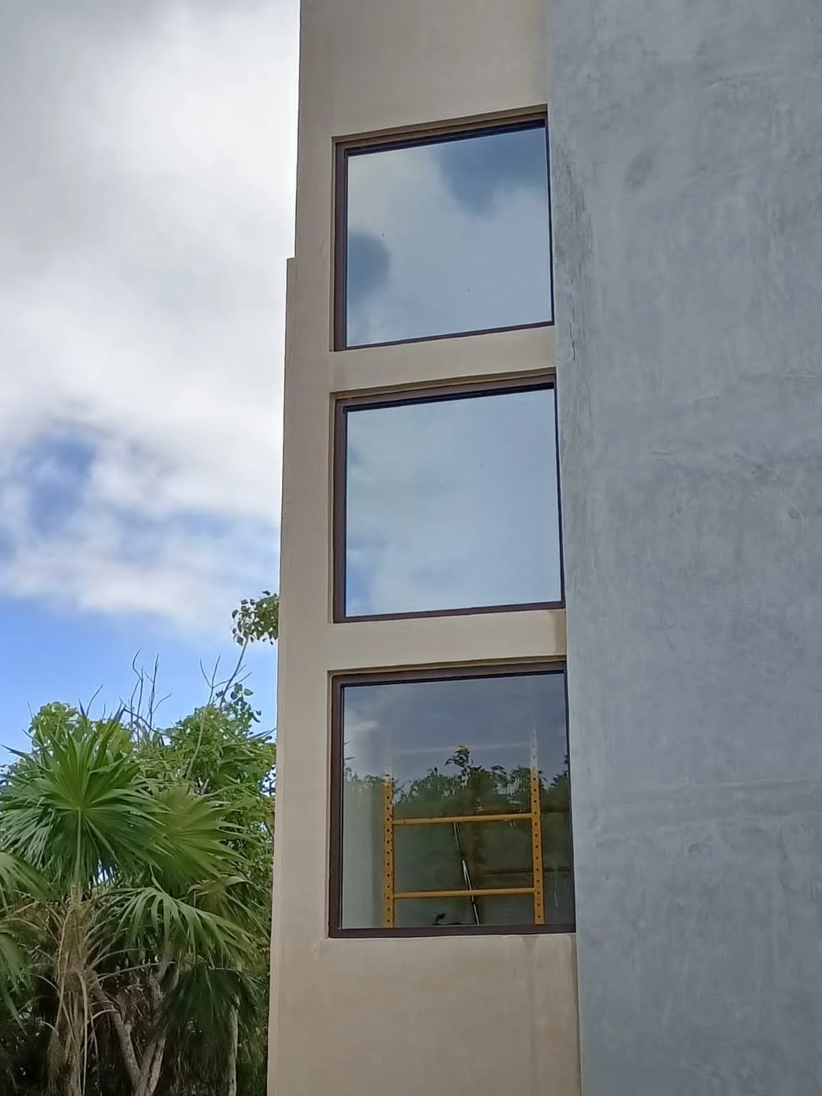 The image shows a modern building with three large rectangular windows, a partly cloudy sky reflected in the windows, some green trees, and a yellow ladder inside the building.