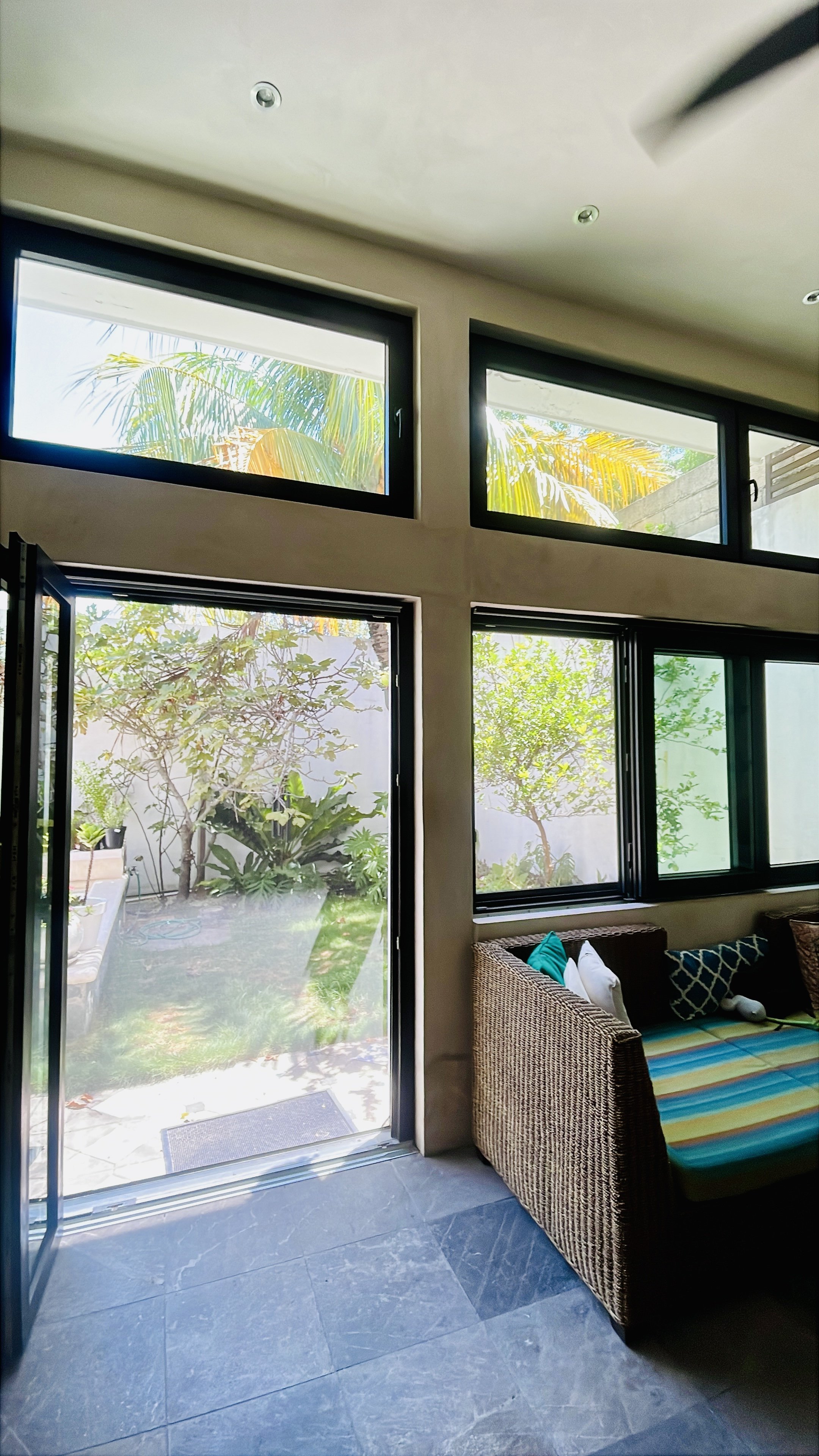 Living room with large windows looking out to a garden featuring trees and green foliage, wicker sofa with colorful pillows, gray tiled floor, and partly visible ceiling lights.