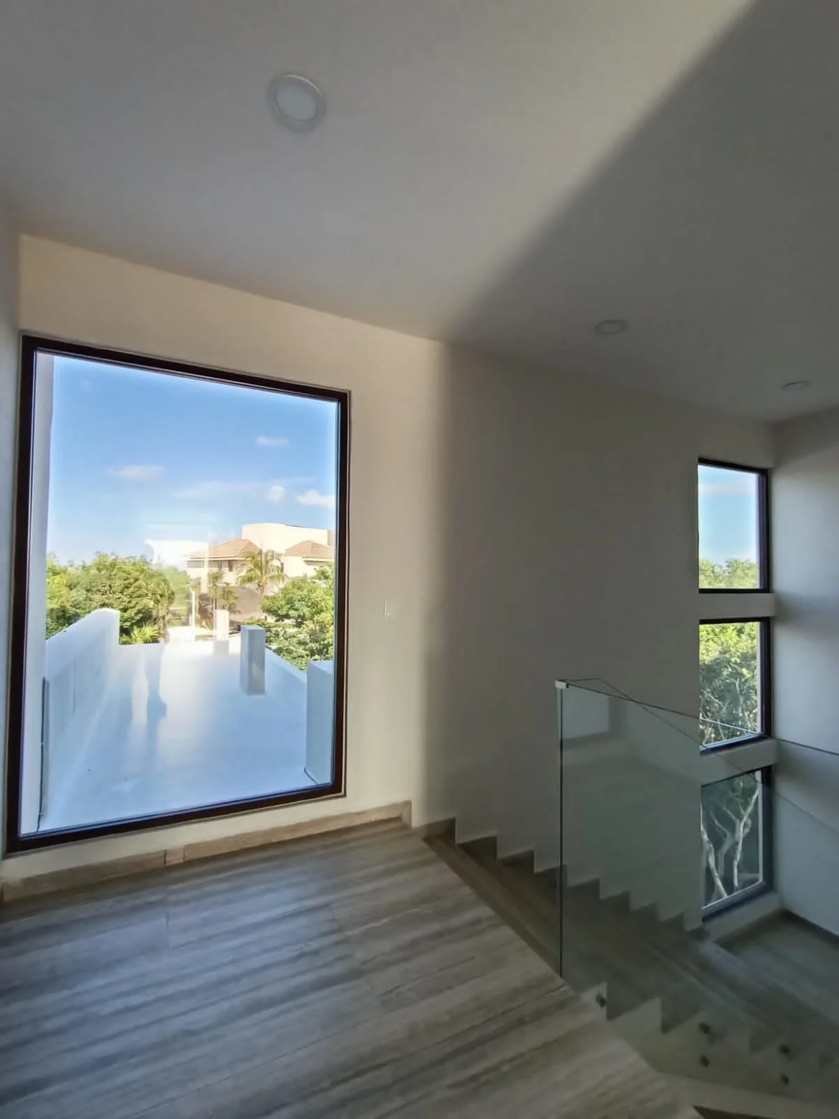 Interior view of a modern home featuring large windows, a staircase with glass railing, and wood-patterned flooring, with a view of trees and neighboring houses outside.
