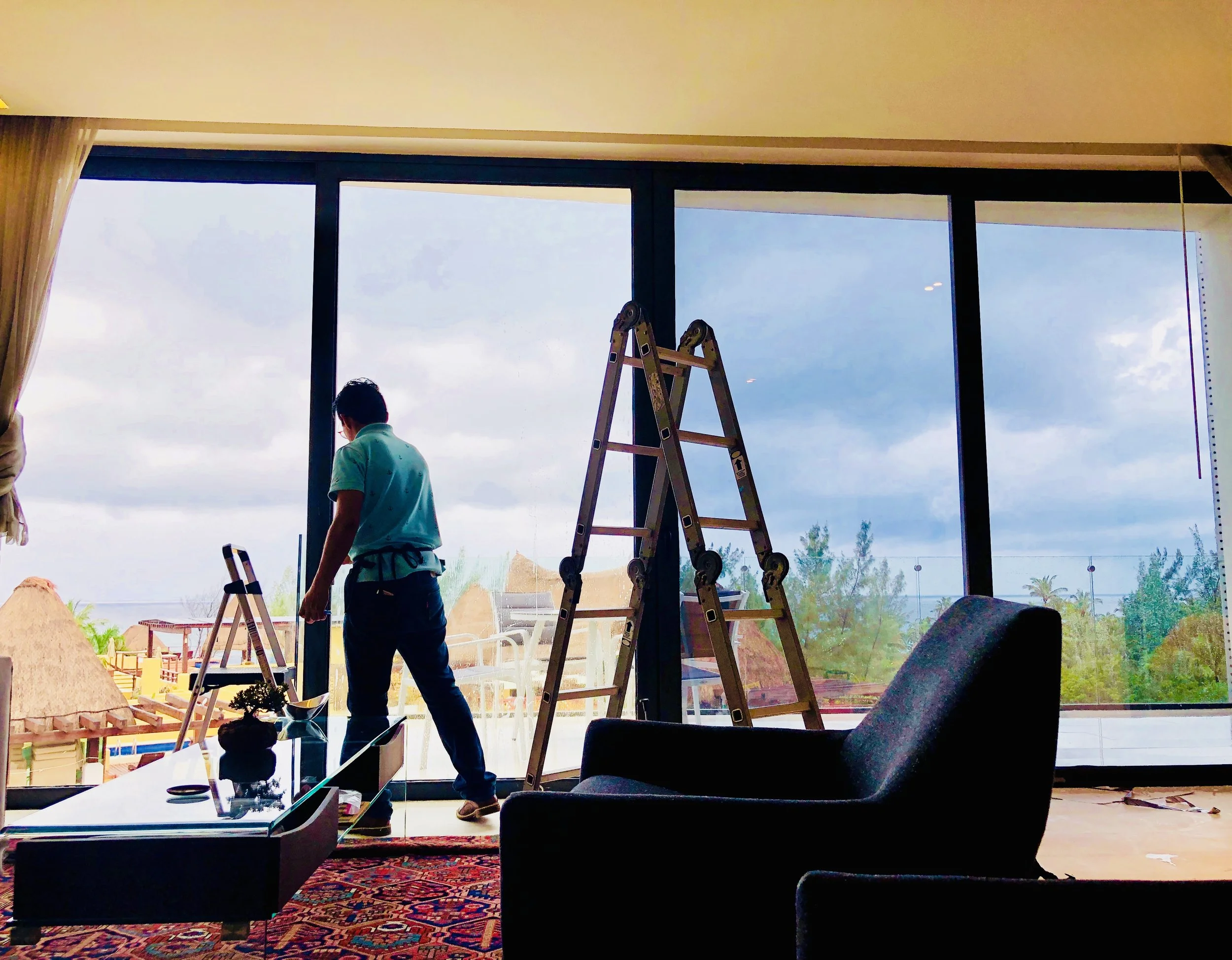 People working on construction or renovation inside a building, with large windows showing an outdoor view of sky and buildings, ladder, and construction materials present.