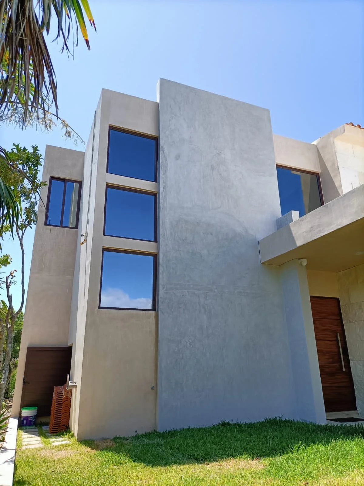 Modern multi-story house with beige exterior, large blue-tinted windows, and a small lawn in the foreground, trees to the left, and a blue sky.