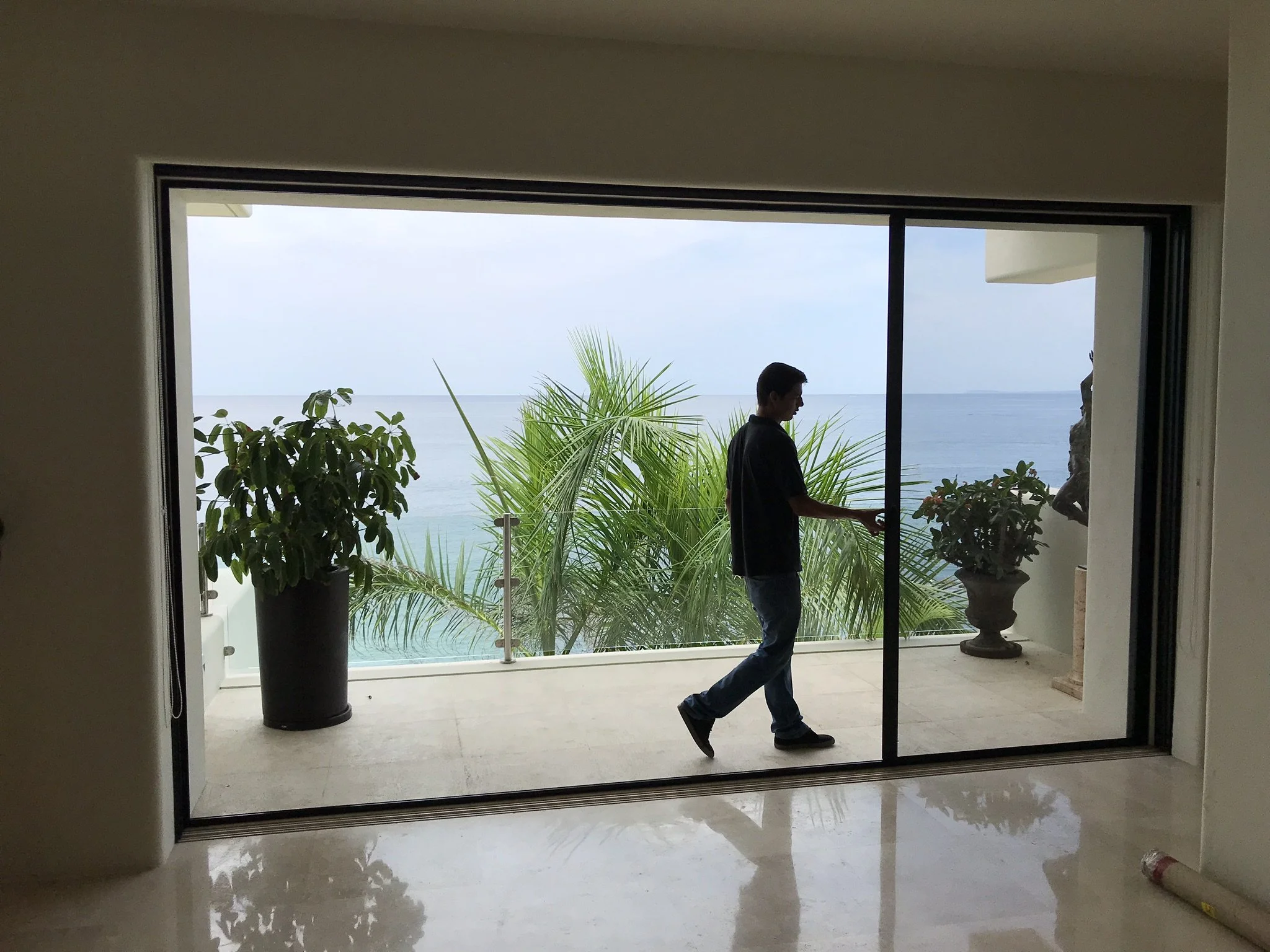 Interior view of a room with a large sliding glass door opening to a balcony with plants, overlooking the ocean, a man is walking in the room.