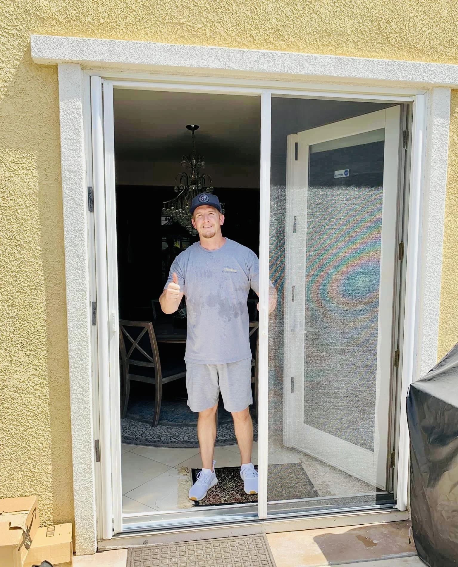 A man standing at a sliding glass door, smiling and giving a thumbs-up gesture, wearing a baseball cap, t-shirt, shorts, and sneakers inside a house.