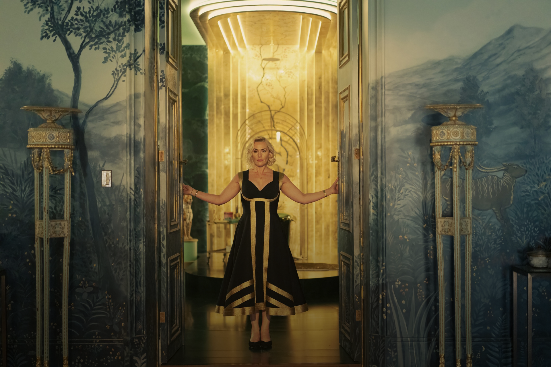 A woman in a black and gold dress is standing in a doorway with intricate gold details, leading to a warmly lit hall with ornate décor and a painted blue and green landscape mural on the walls.