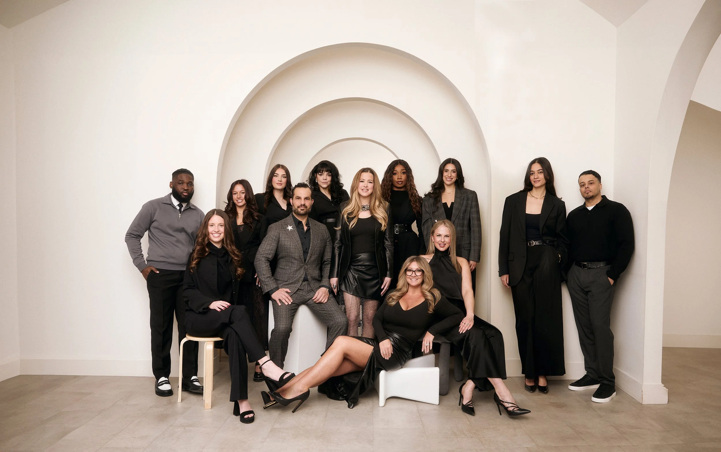 Group of 12 diverse people, mostly women in black attire, posing in a modern minimalist indoor setting with a fluted arch backdrop.