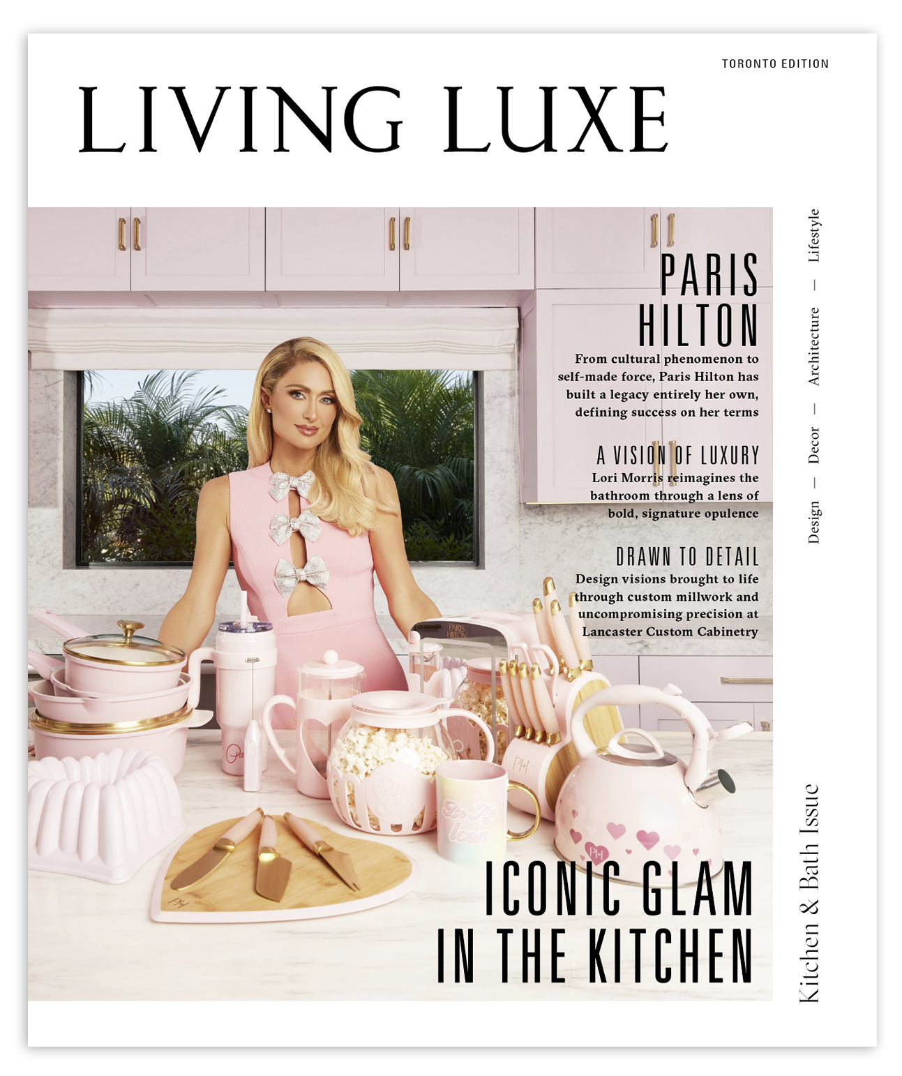 A woman with blonde hair in a pink dress with bow details is standing in a kitchen surrounded by pink kitchenware including a teapot, mugs, a popcorn bowl, knives, and a cutting board. Behind her, there is a window with green tropical plants outside, and white cabinets above.
