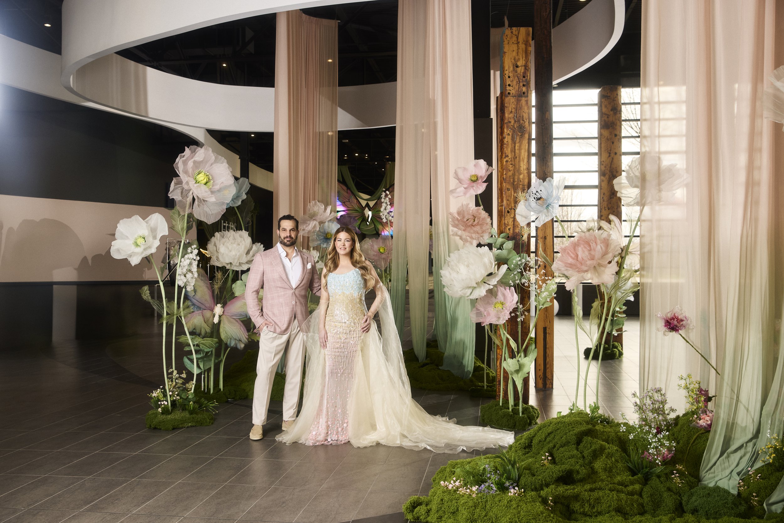 A couple standing together in front of a floral and fabric backdrop at a wedding or event, with large flowers and greenery surrounding them.