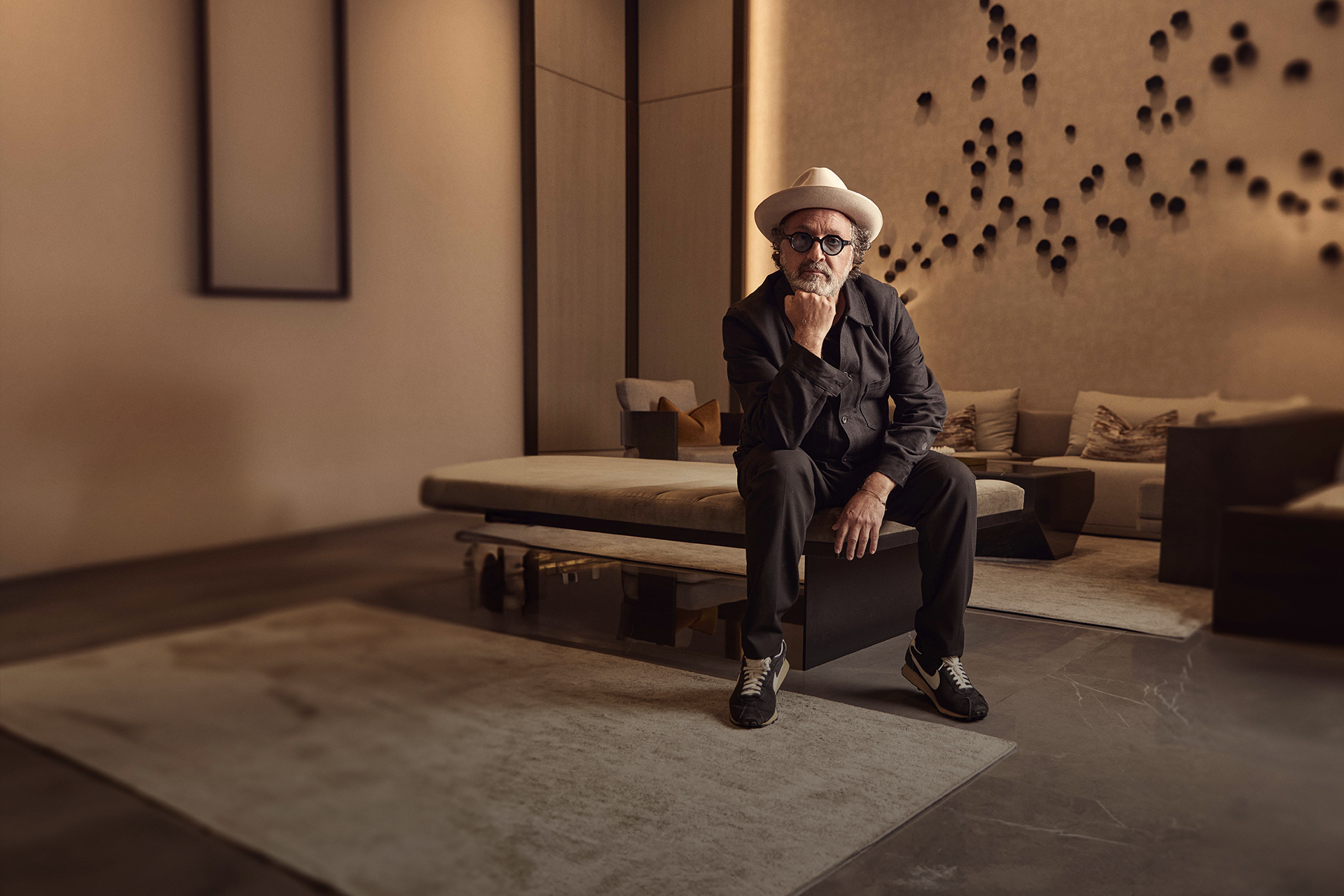 A man with glasses, a beard, and a white hat sits on a bench in a modern, warmly lit room with beige walls and a patterned accent wall. He is dressed in black and wearing sneakers, with one hand resting on his chin and his other arm resting on his knee.