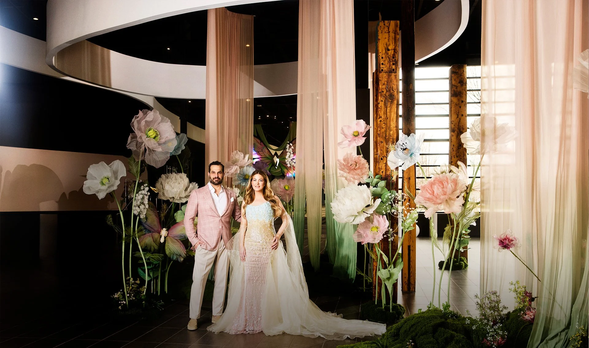 A couple stands in a floral archway inside a modern building. The man wears a light pink plaid suit with a white shirt and no tie and has a beard. The woman wears a light-colored, elegant dress with a sheer overlay and long train, and has long wavy hair. Large decorative flowers and vertical fabric drapes surround them.