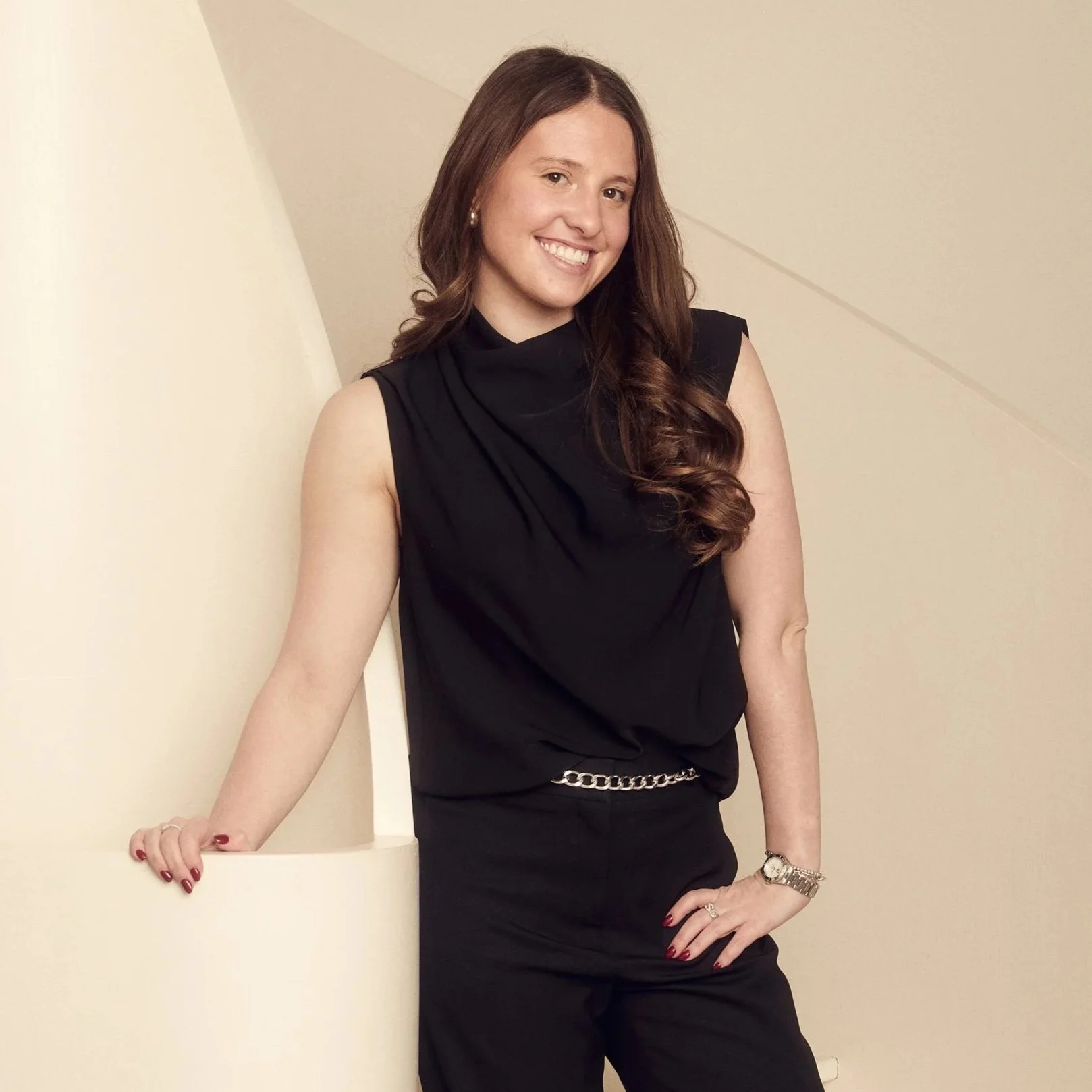A woman with long wavy brown hair wearing a black sleeveless top and black pants, standing against a light-colored wall, smiling with her left hand on her hip, accessorized with a gold watch, bracelet, and ring.