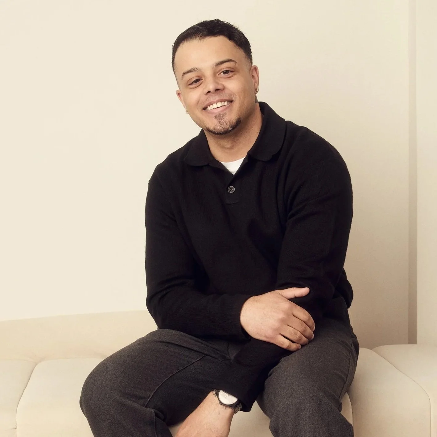 A man sitting on a cream-colored sofa, smiling at the camera. He has short dark hair, a goatee, and is wearing a black long-sleeve shirt and black pants.