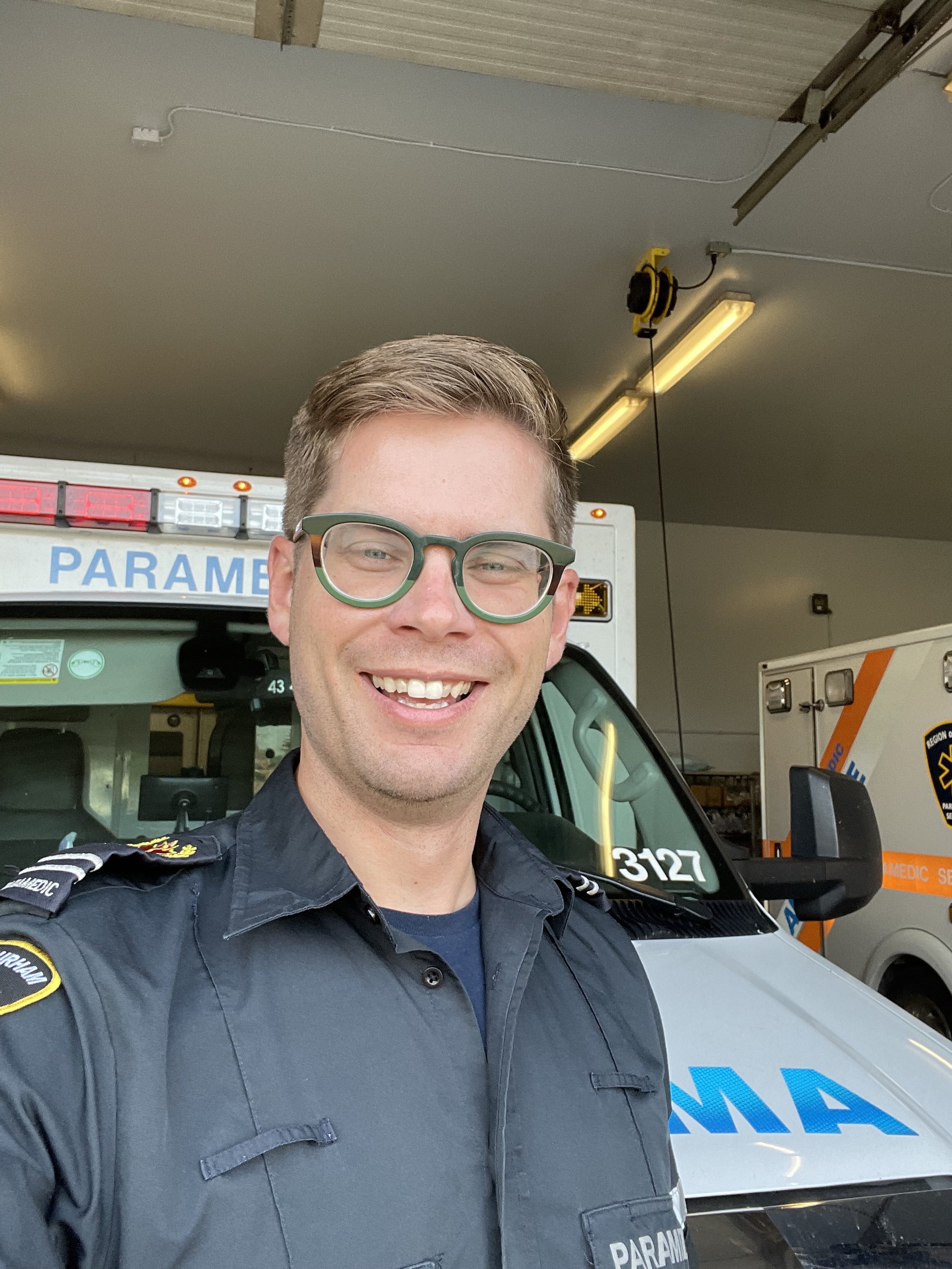 A smiling paramedic taking a selfie in front of an ambulance inside a garage.