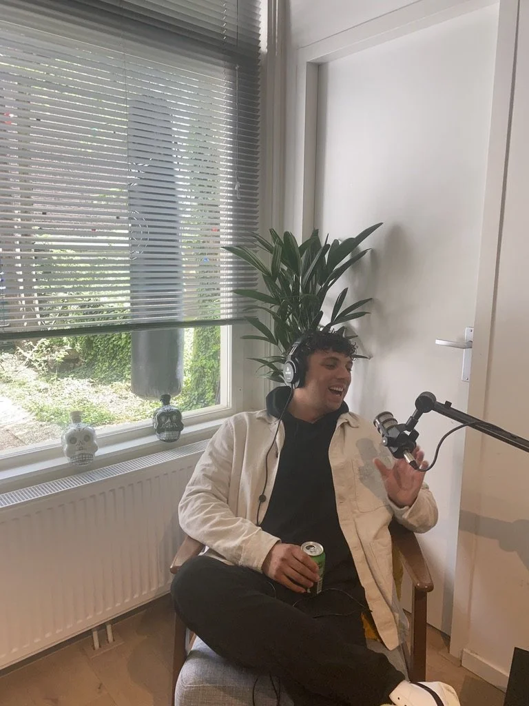 Man die in een stoel zit, muziek luistert via koptelefoon, lachend met een blik op de microfoon voor een opname, in een kamer met een raam, decoratie schedels en een grote plant.