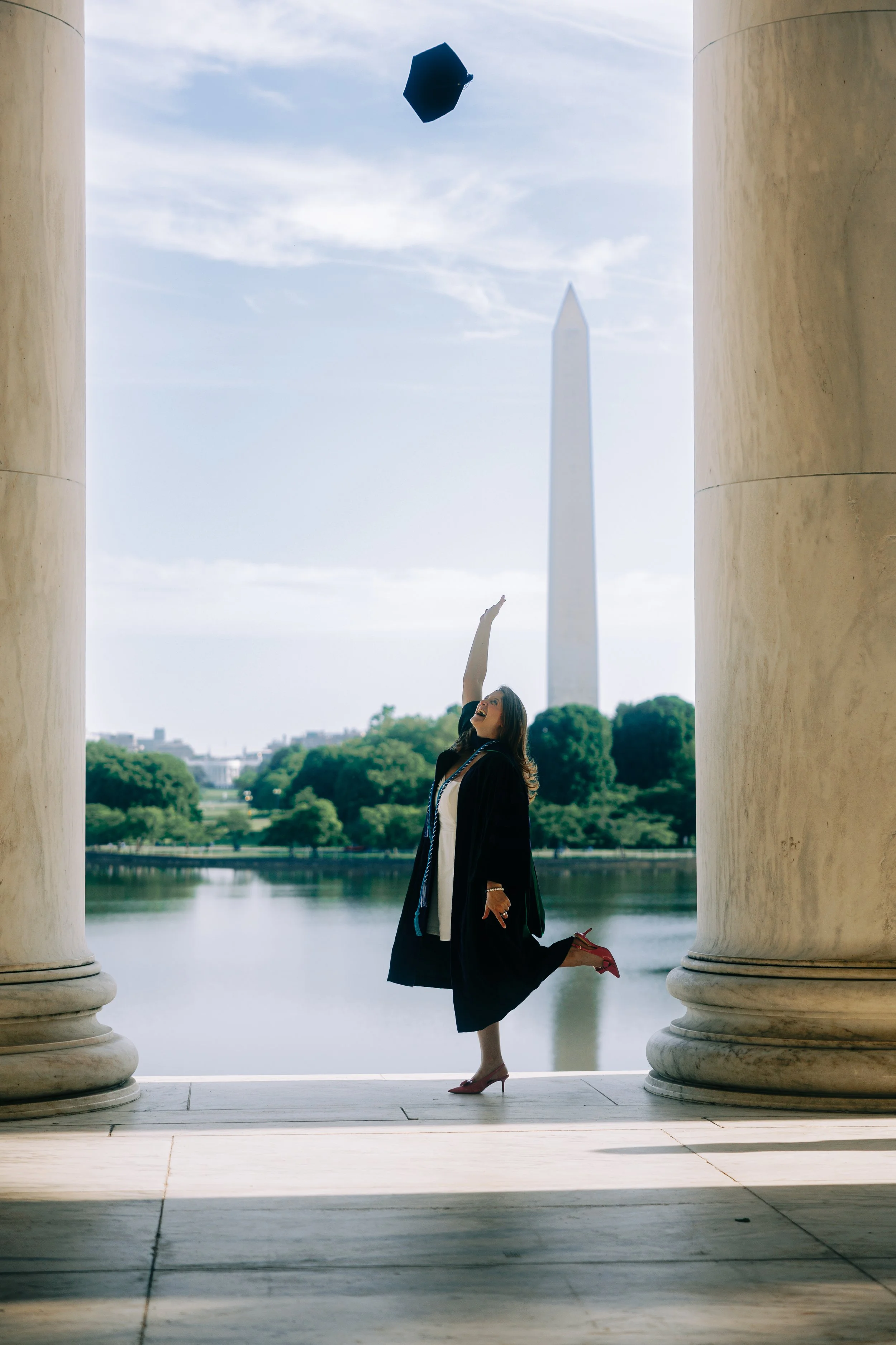 A woman in a graduation gown and cap jumping into the air with the Washington Monument in the background in Washington, D.C.