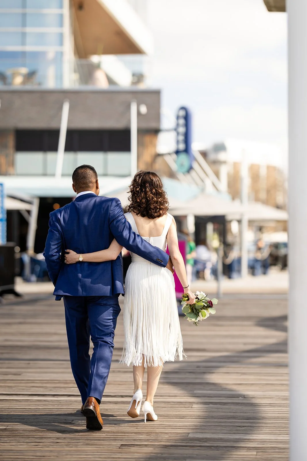 A couple walking on a wooden boardwalk, with the man in a blue suit and the woman in a white dress holding a bouquet, seen from the back.
