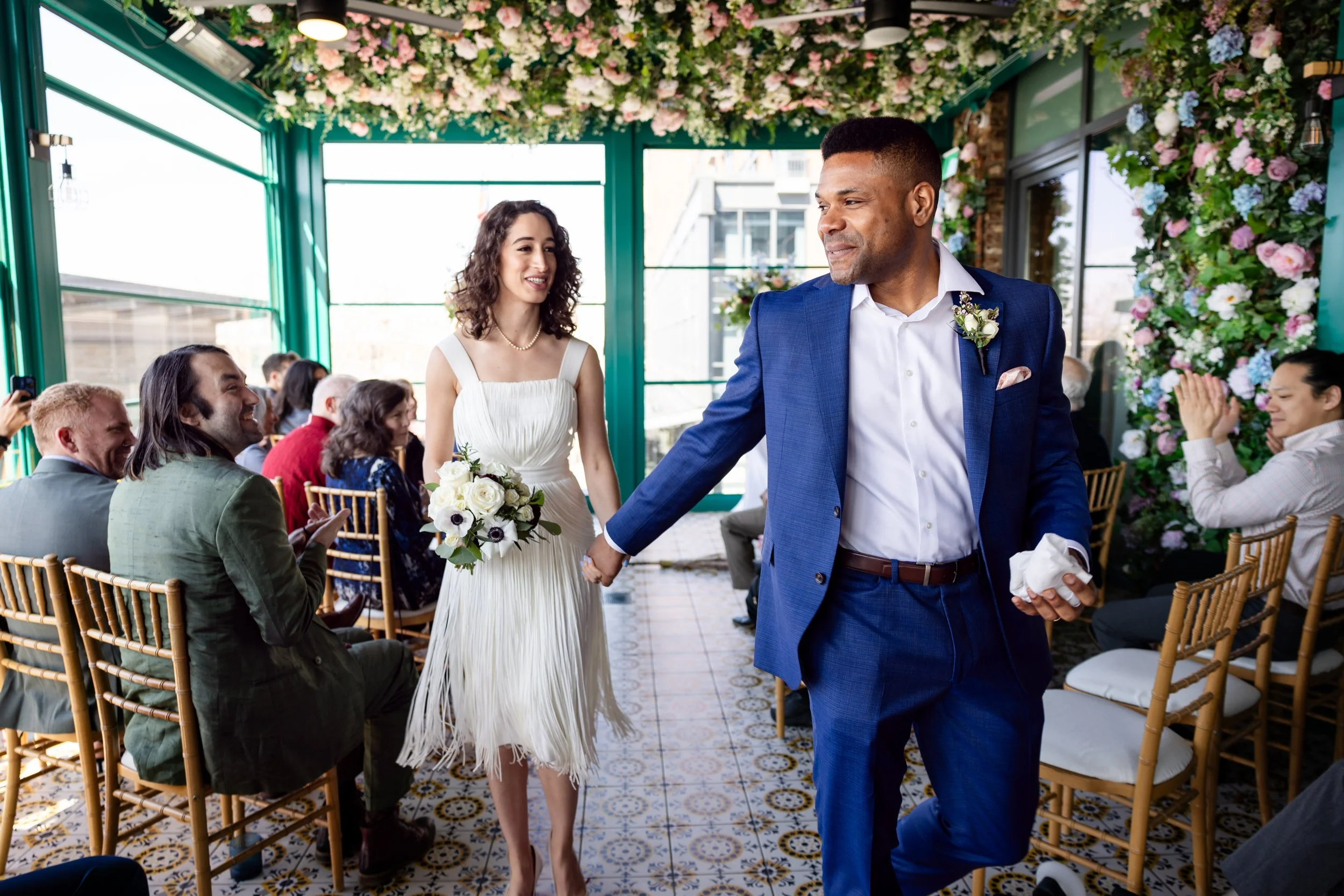 A wedding reception with a bride and groom holding hands, walking through guests seated at tables. The bride wears a white dress and holds a bouquet, and the groom is dressed in a blue suit. The room is decorated with flowers on the ceiling and walls