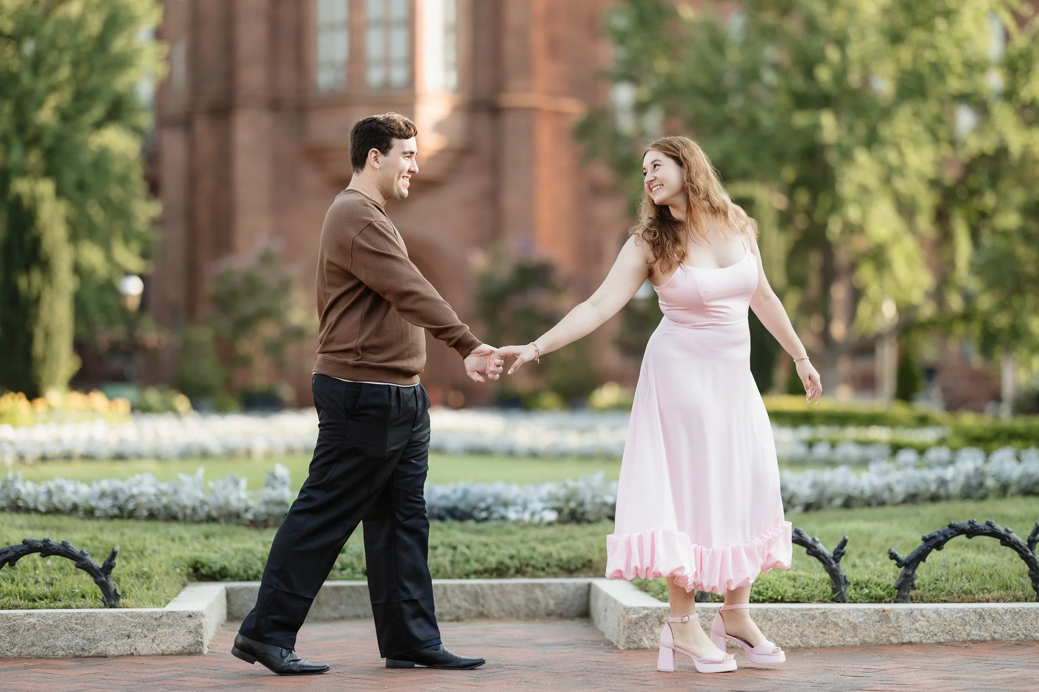 A man and woman are holding hands and smiling at each other in a park with a brick building and trees in the background. The woman is wearing a pink dress and pink high heels, and the man is dressed in a brown sweater and black pants.